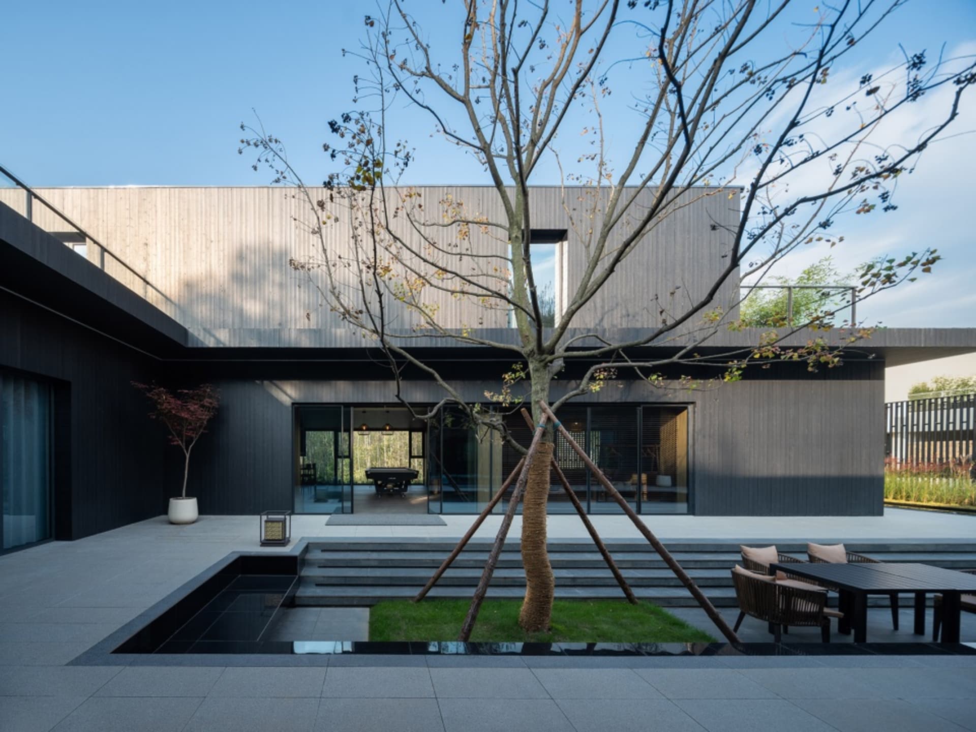 A modern house with black walls, a glass door, and a tree in front, featuring two chairs and a dining table outside