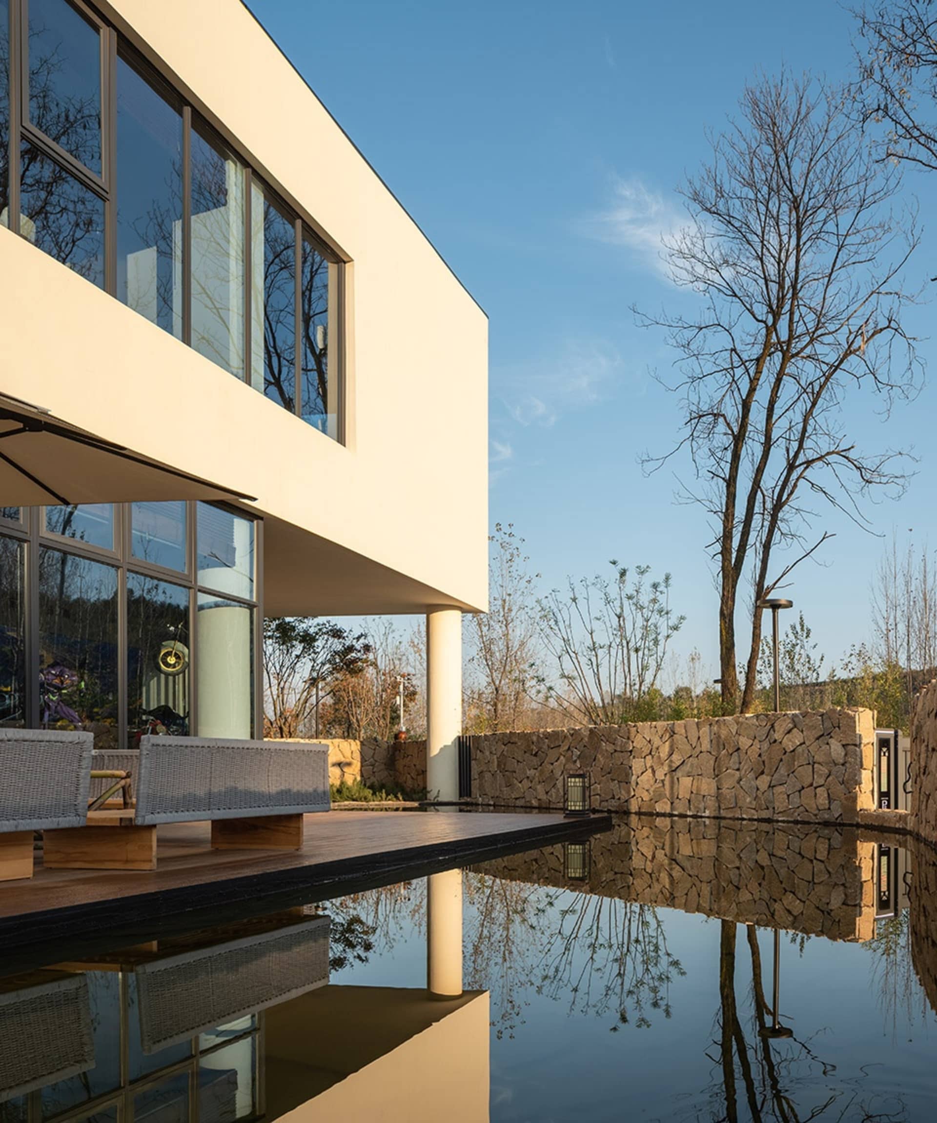 Modern house with a large glass wall overlooking a pond, featuring a living area with couches and chairs, and an exterior made of concrete