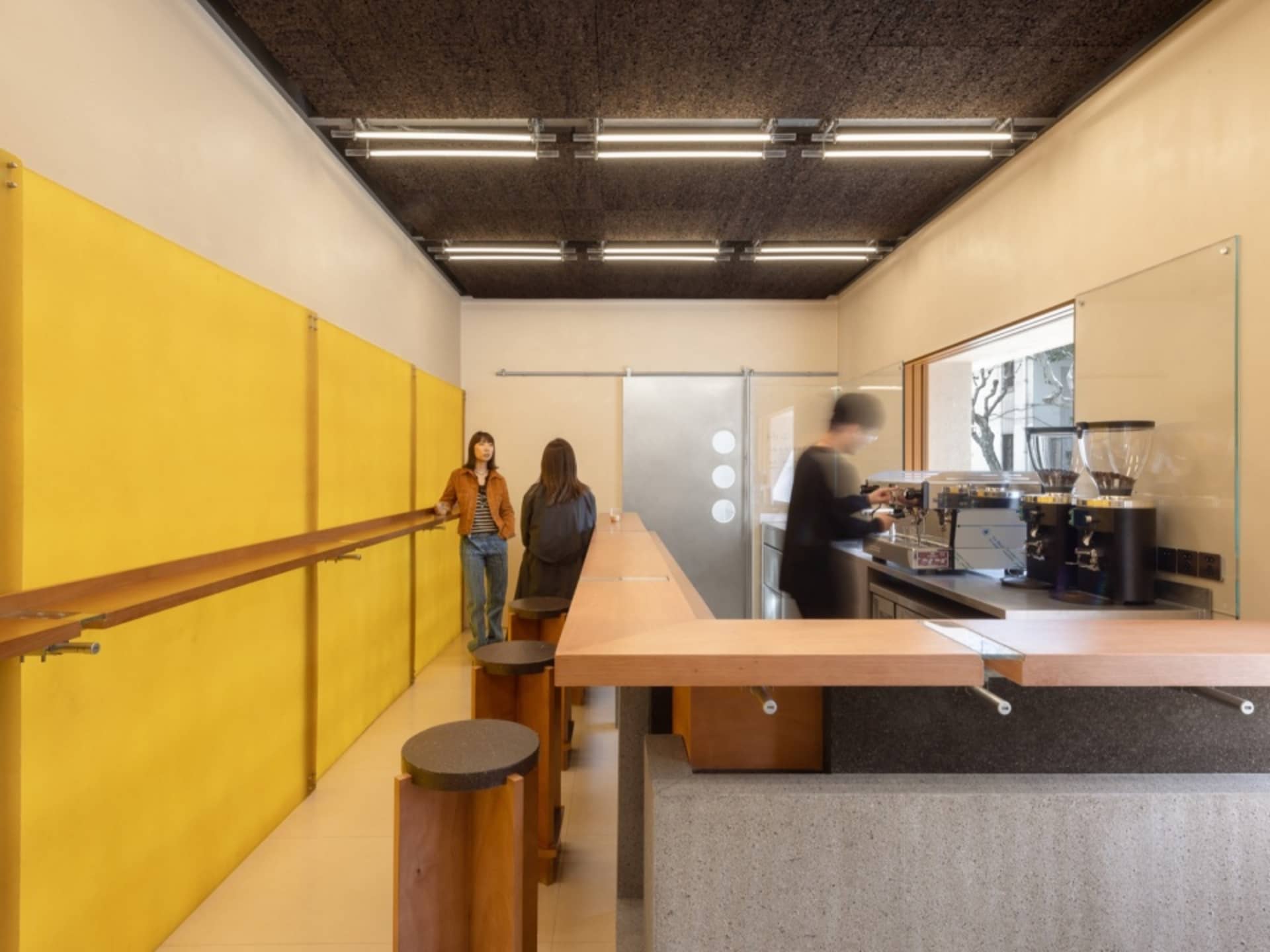 A modern restaurant with a counter and stools, featuring wooden surfaces and metal accents, warm lighting, and three women engaged in conversation