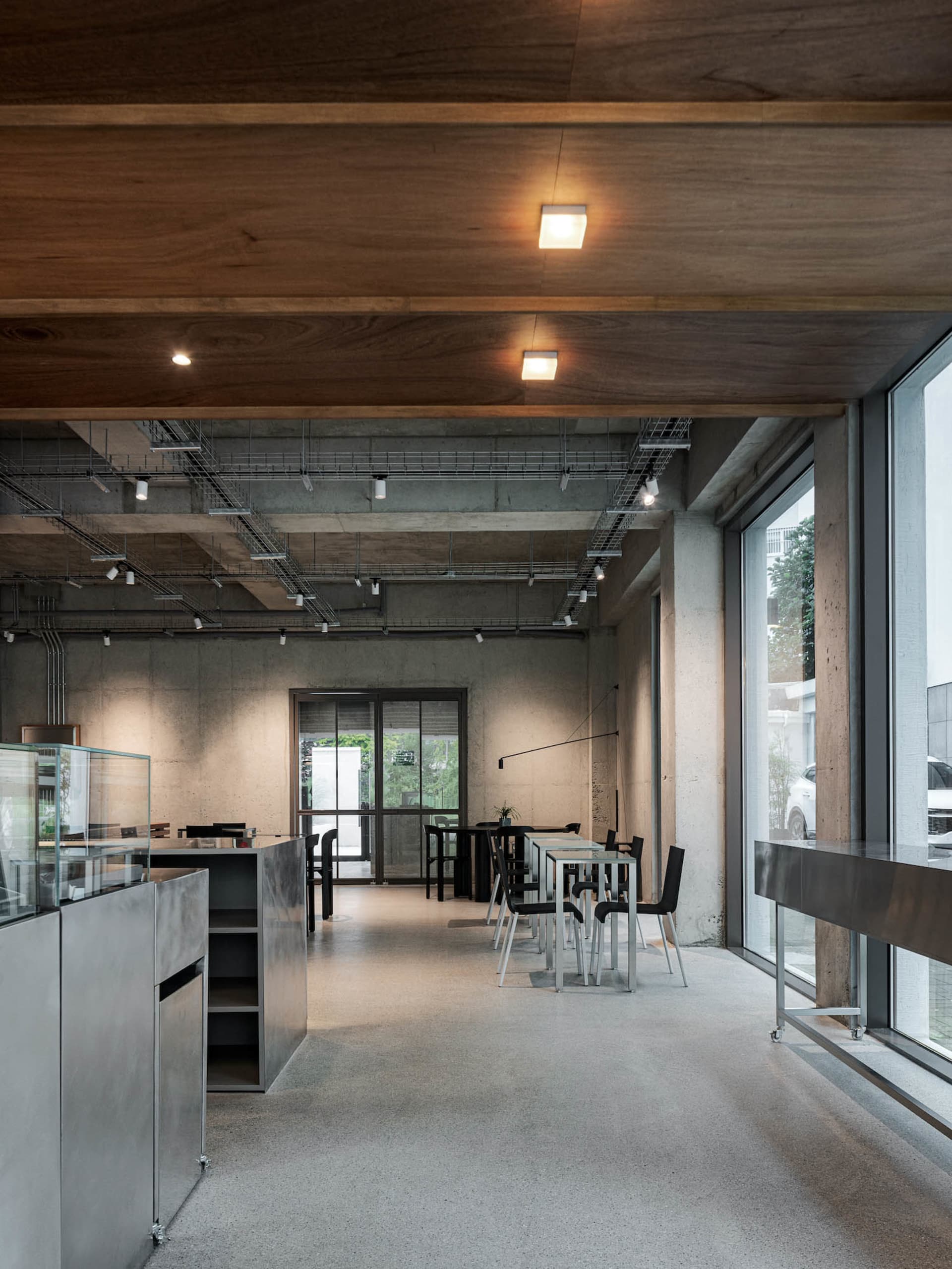 Modern restaurant interior with multiple dining tables, chairs, concrete walls, and wooden accents, illuminated by natural light
