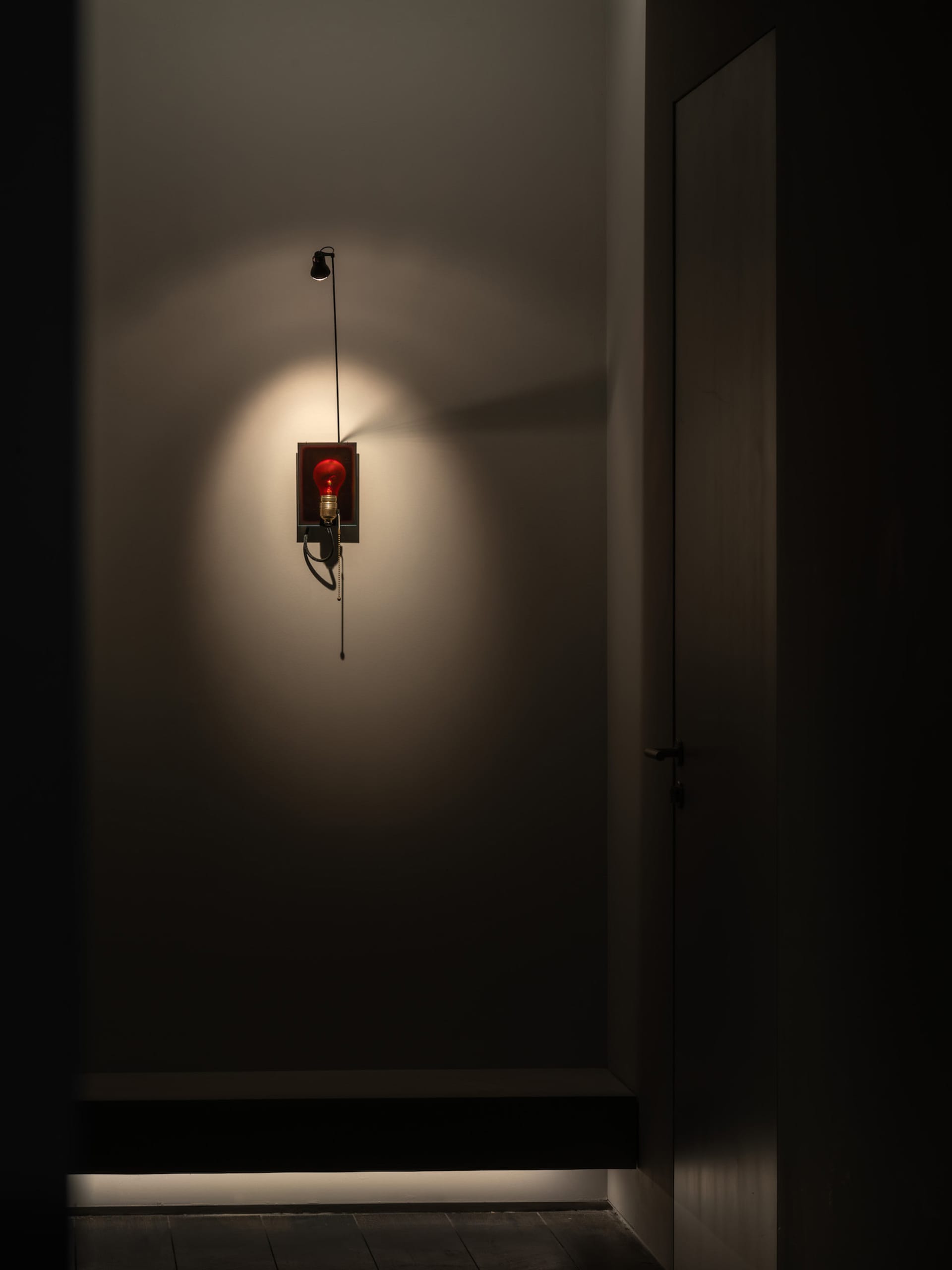 A dimly lit hallway with a door and a fire alarm with a red light, mounted to the wall