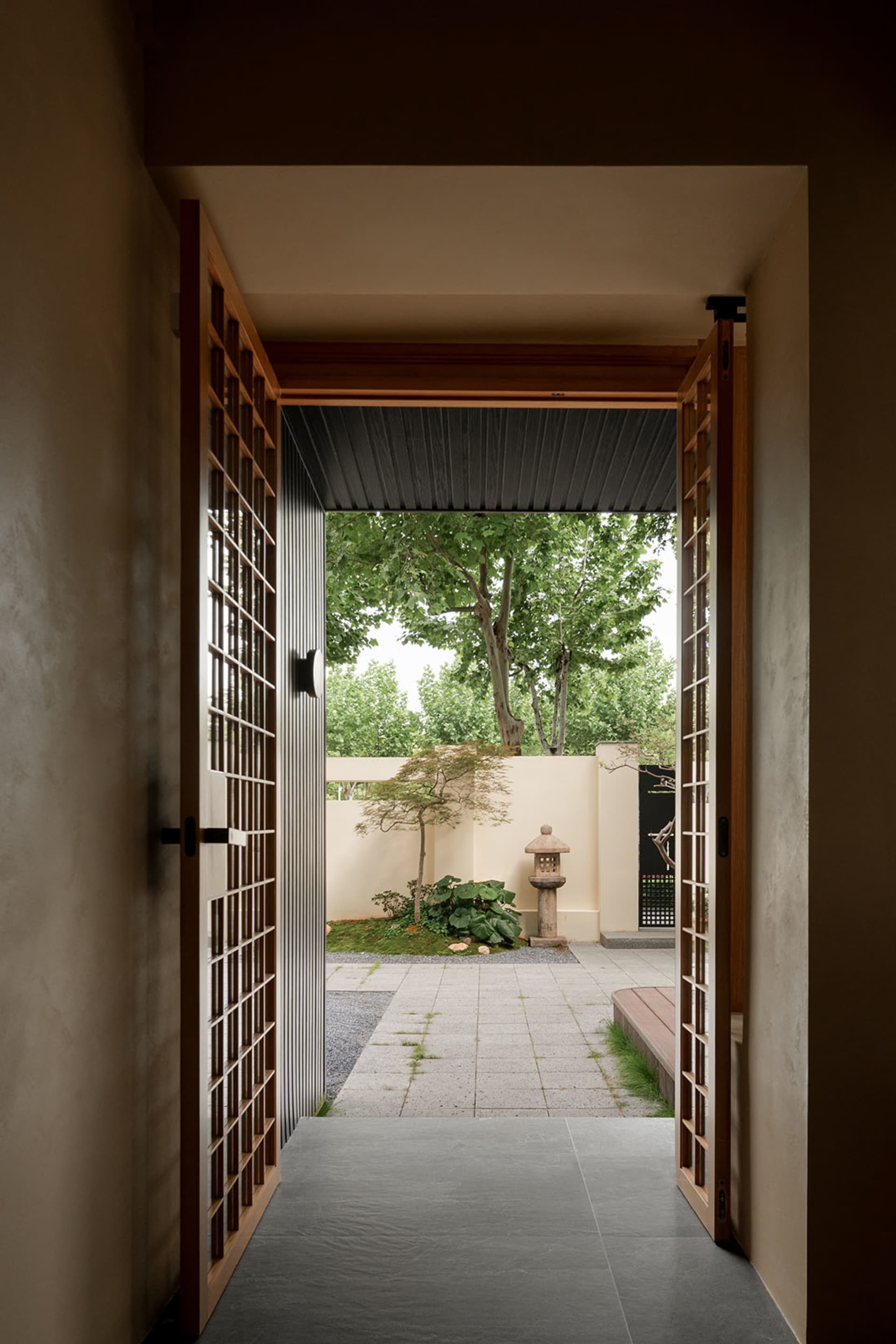 A serene outdoor entrance with a wooden-framed glass doorway, surrounded by two trees and a calm atmosphere