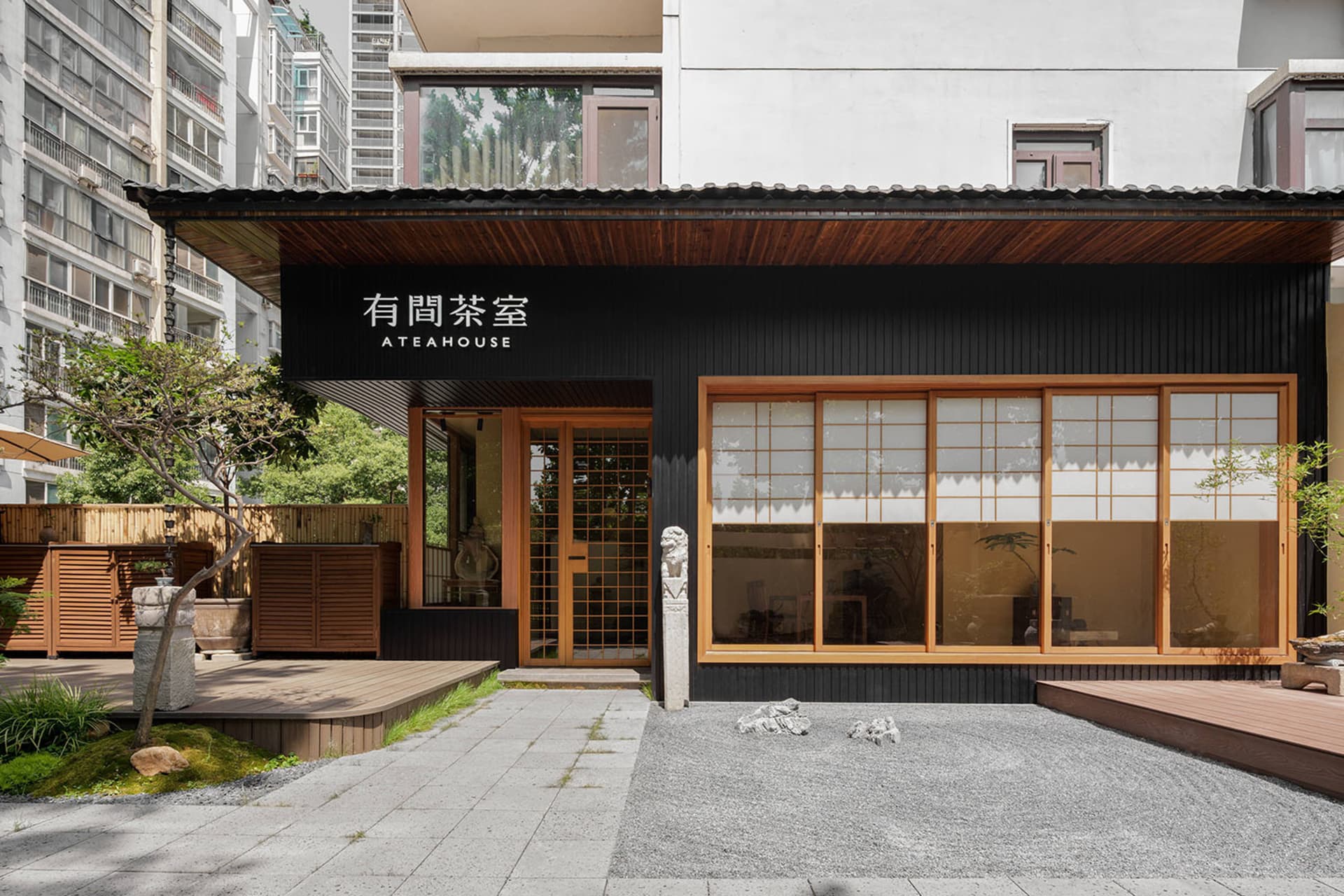 Modern Asian restaurant with a black facade, wooden elements, and natural light from windows, featuring a dining area with tables and chairs and potted plants