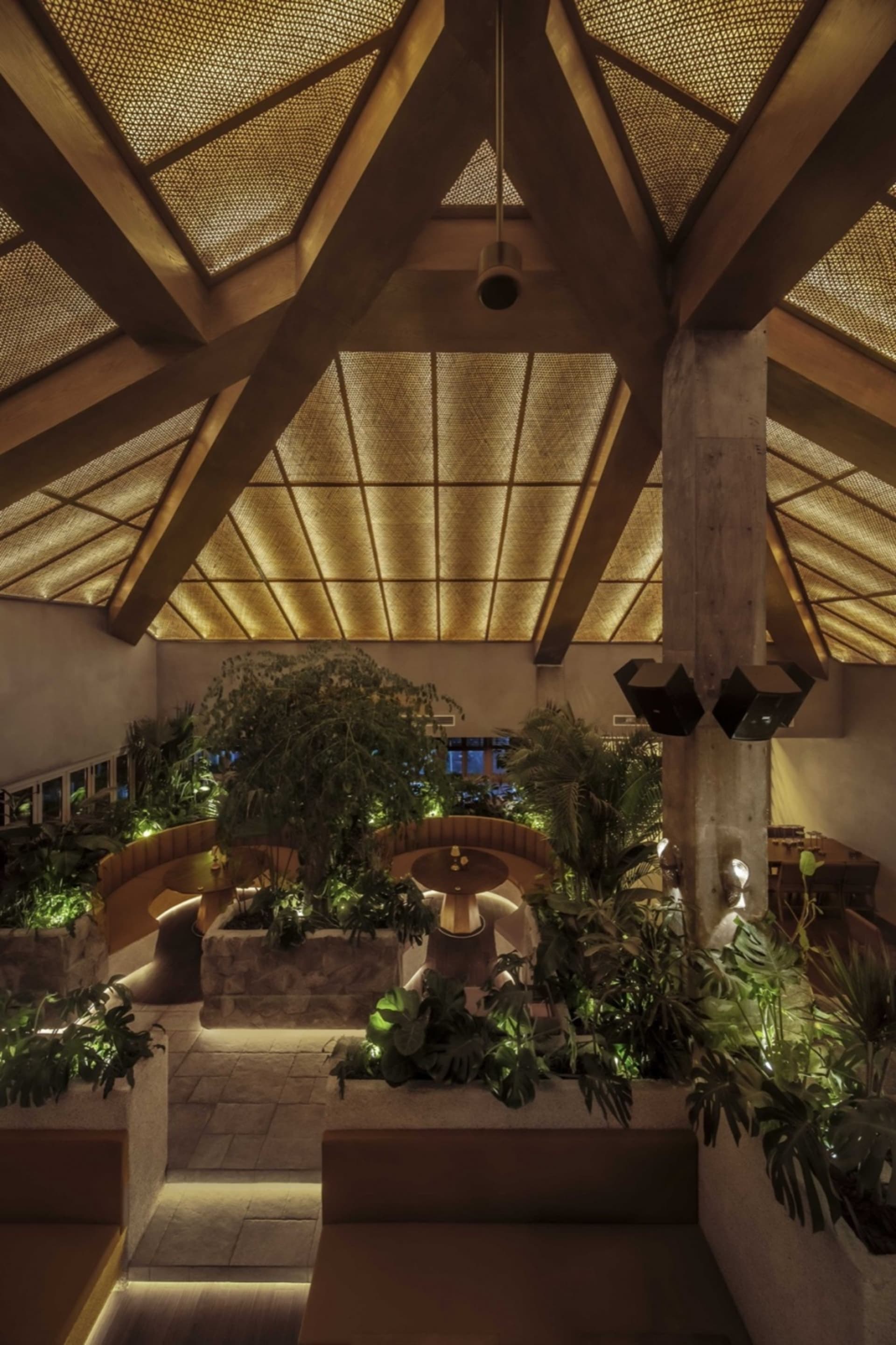 A modern restaurant interior with high ceilings, wooden beams, and stone elements, featuring potted plants and dining tables in a welcoming atmosphere