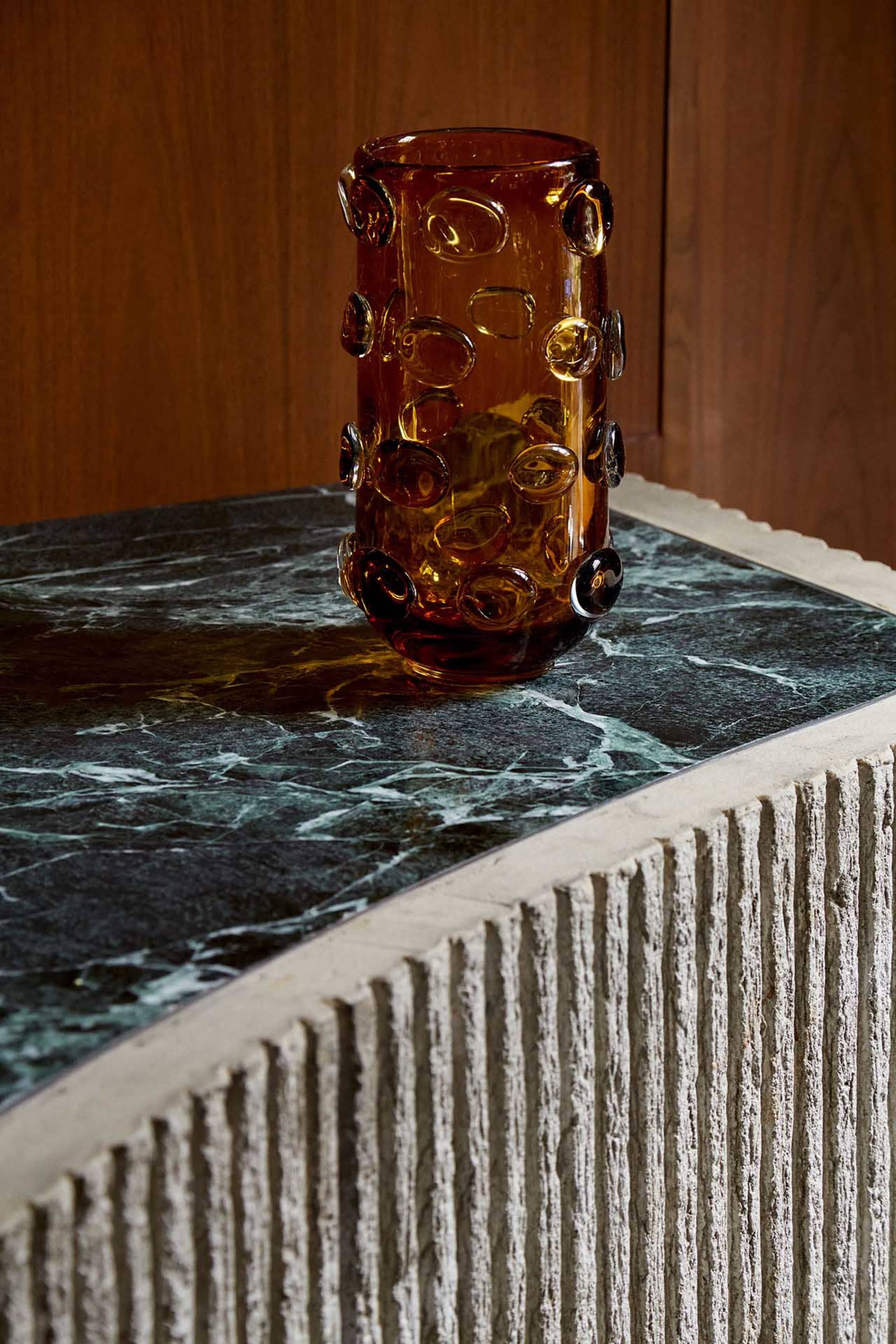 A vase sits on a marble table in front of a wooden wall, creating a sophisticated atmosphere with the intricate design of the vase and the combination of materials