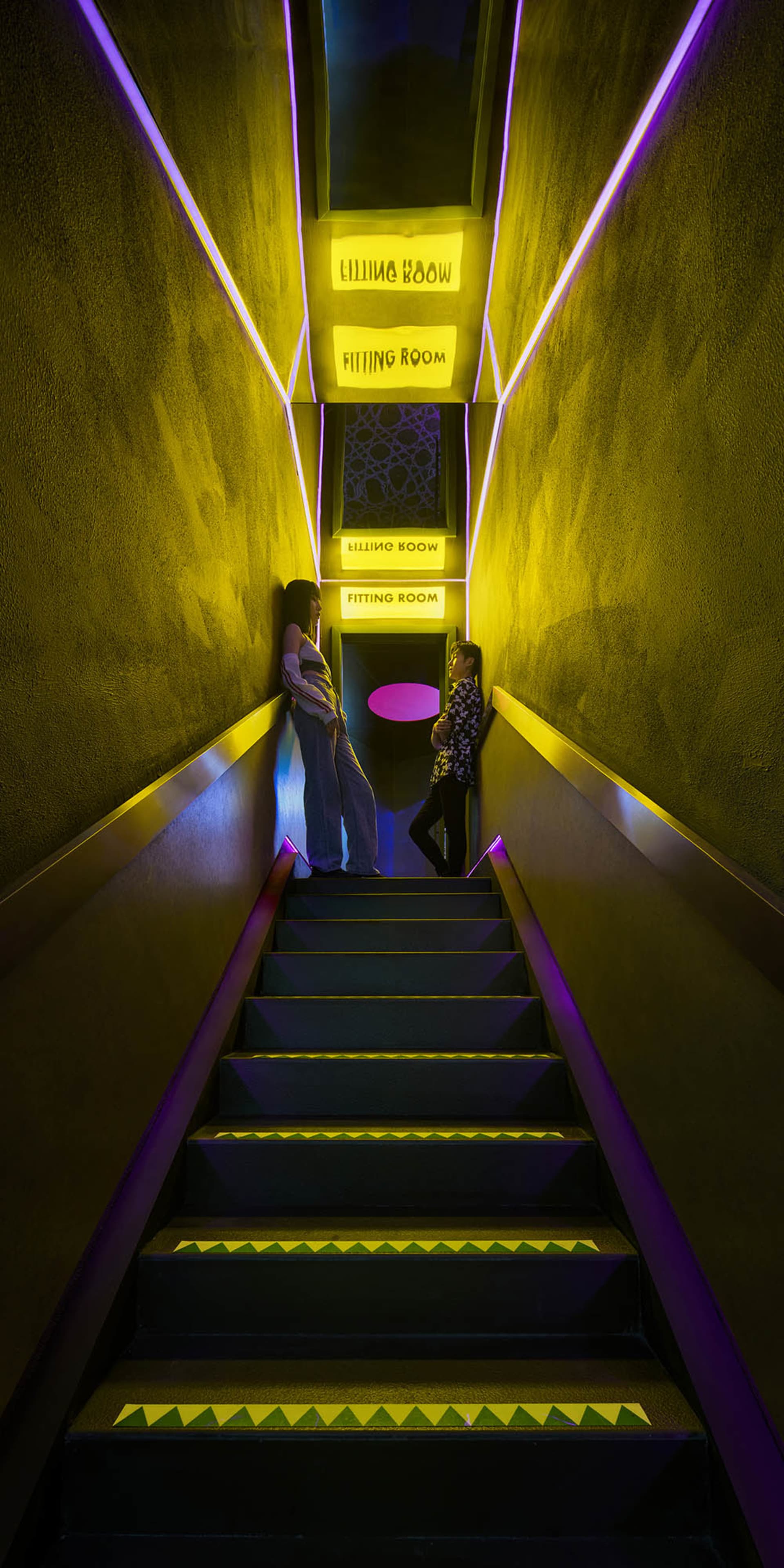 Two women standing on a staircase in a dimly lit room with yellow lights, chairs, and a mix of living and hallway space