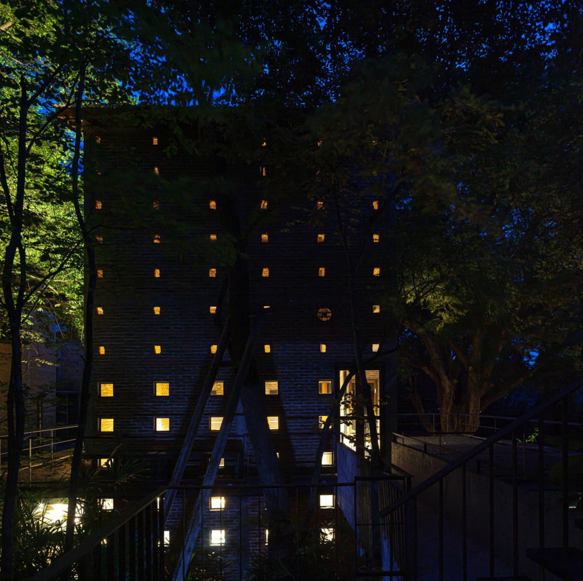 Modern building with a brick exterior, metal staircase, and windows on multiple levels, surrounded by potted plants and captured during dusk