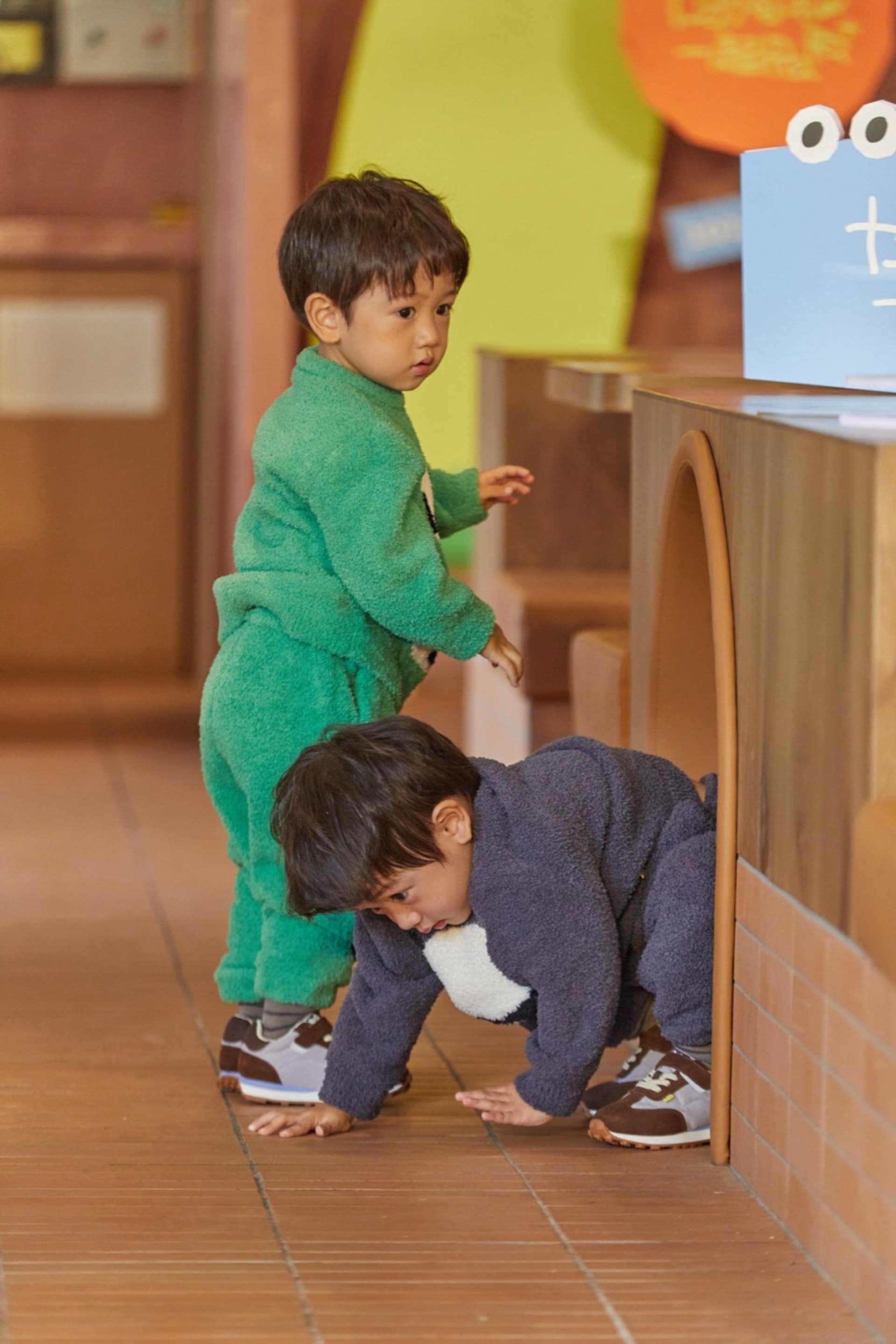 A warm and inviting space with wooden floors and walls, featuring a dining area with chairs and a book, and a young boy playing with a teddy bear on the floor