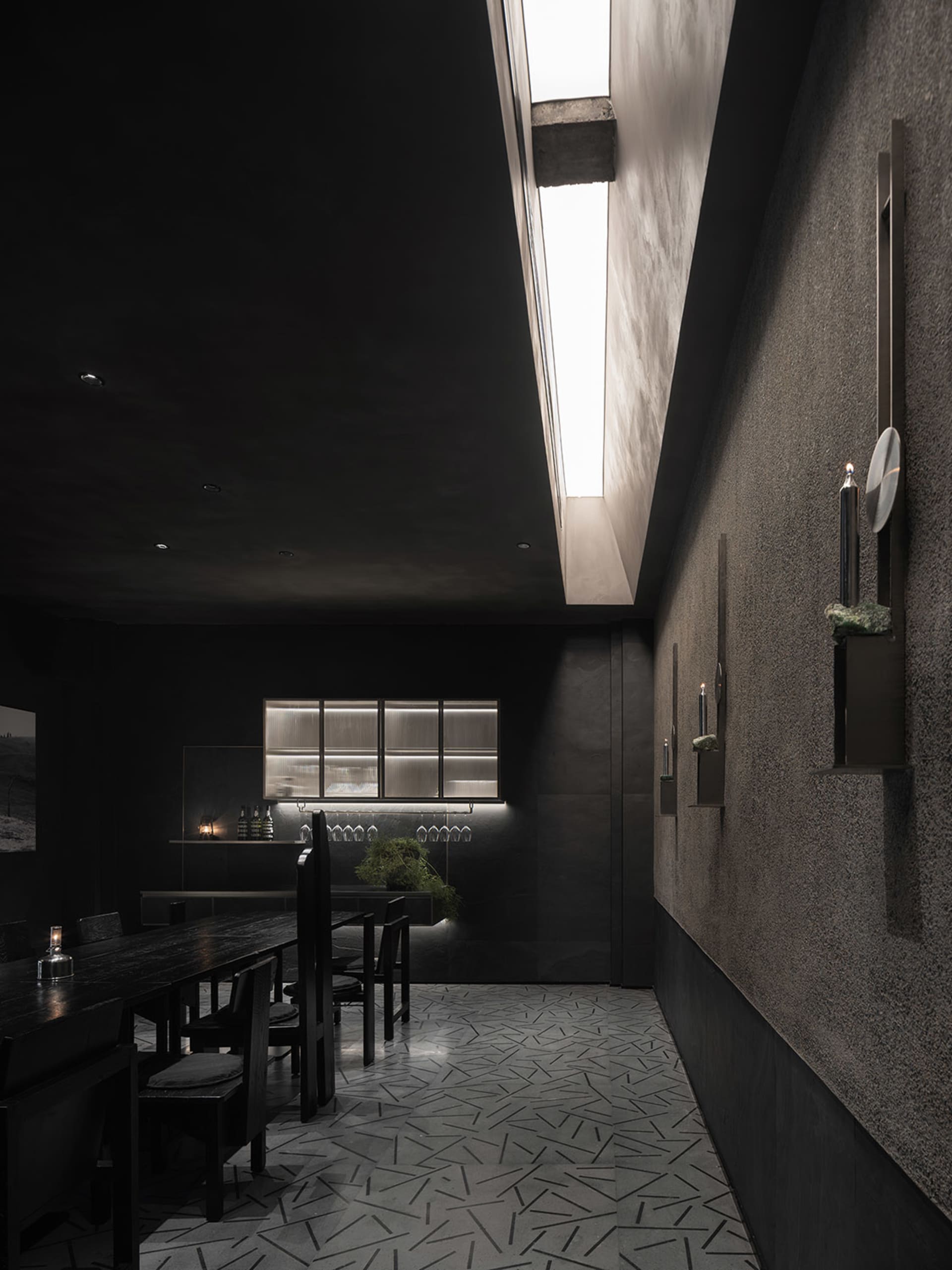 Dark restaurant interior with black walls and floors, featuring dining tables and chairs, and scattered bottles