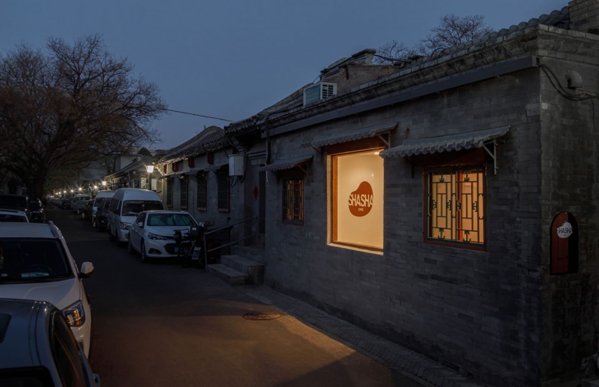 A bustling Asian city street at night with a small brick building, cars, and a truck parked along the street