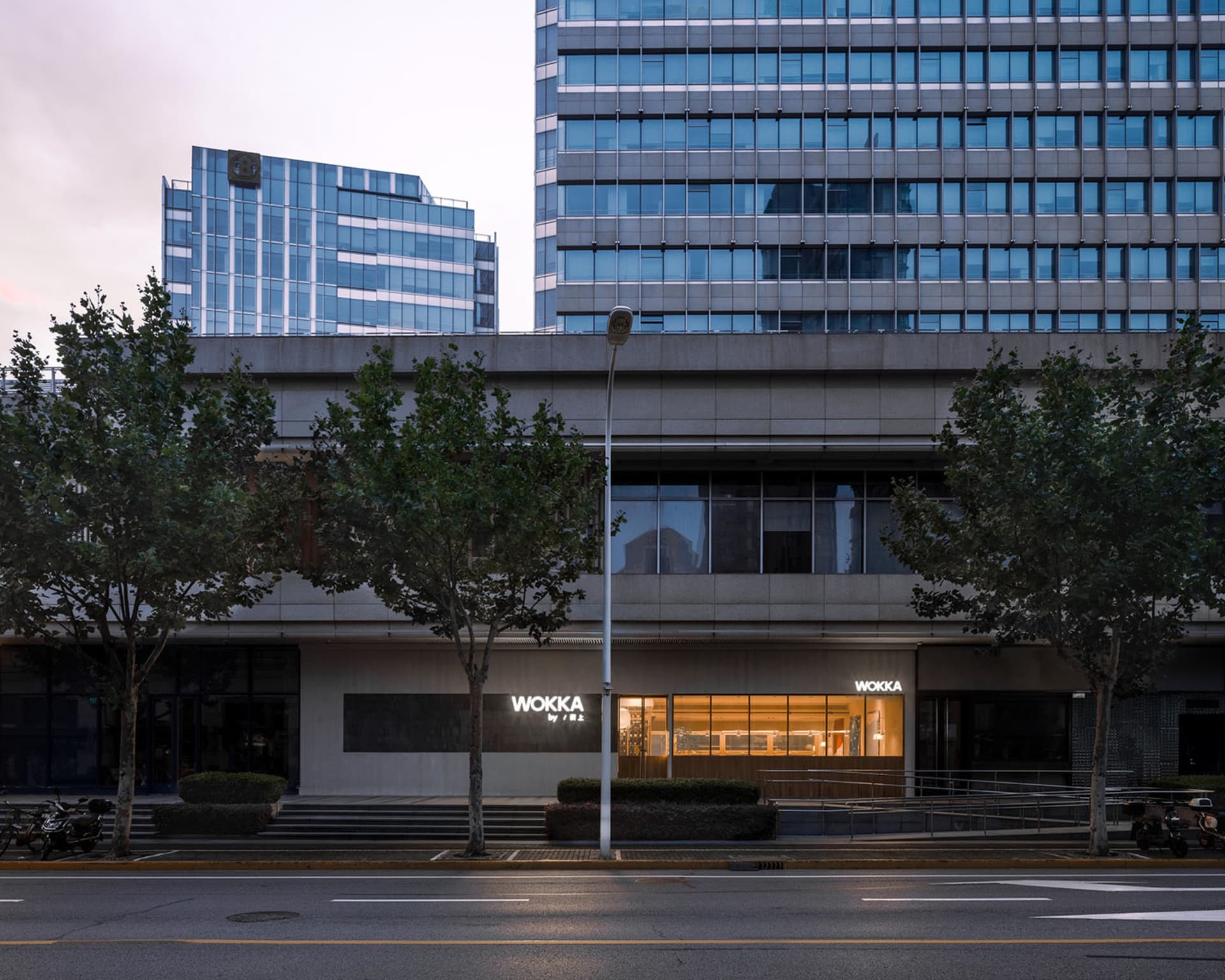 A modern building with multiple floors and glass windows, surrounded by trees and parked cars on a city street