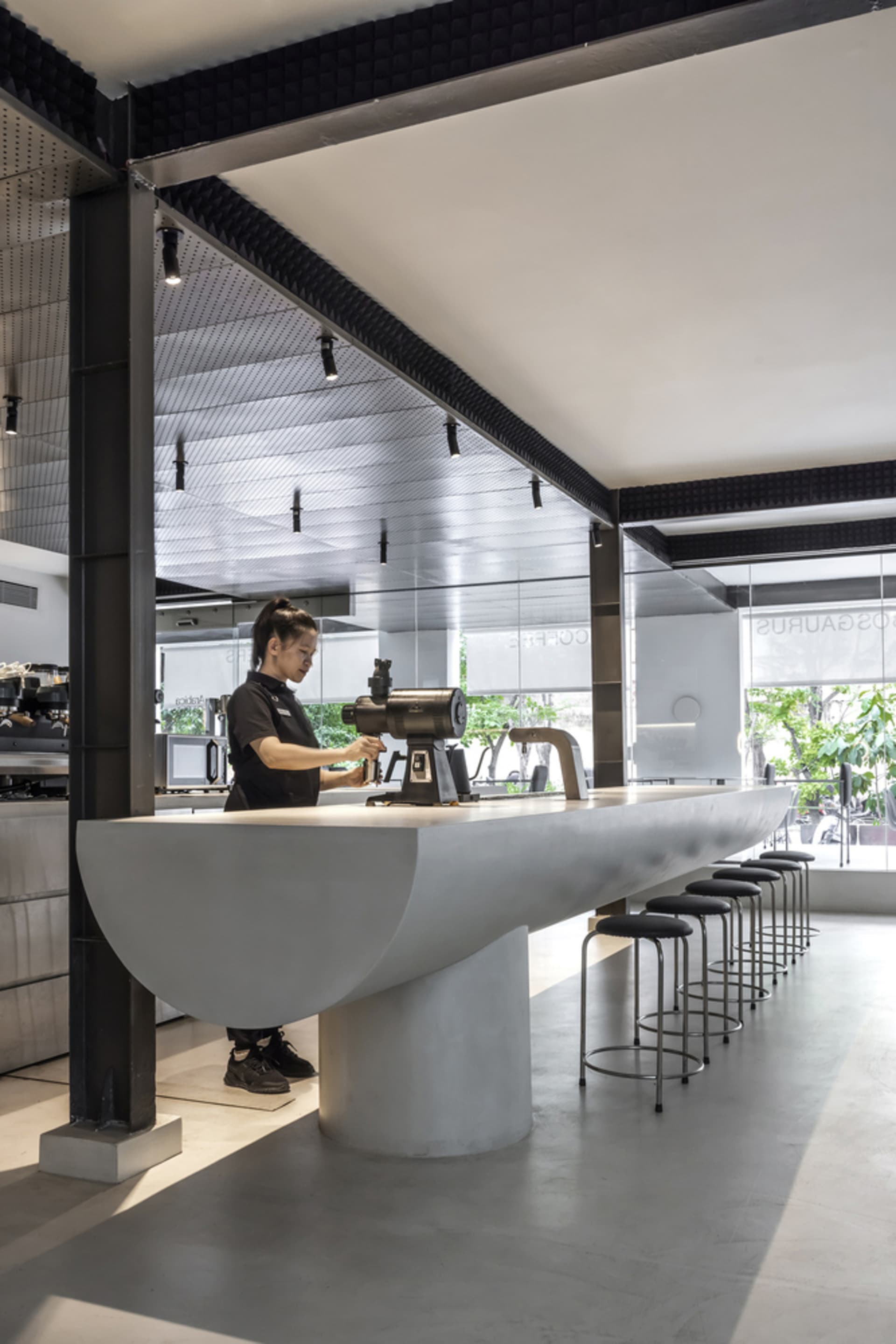 A modern cafe with a long counter and stools, surrounded by chairs and natural light from a large window, with a woman standing behind the counter