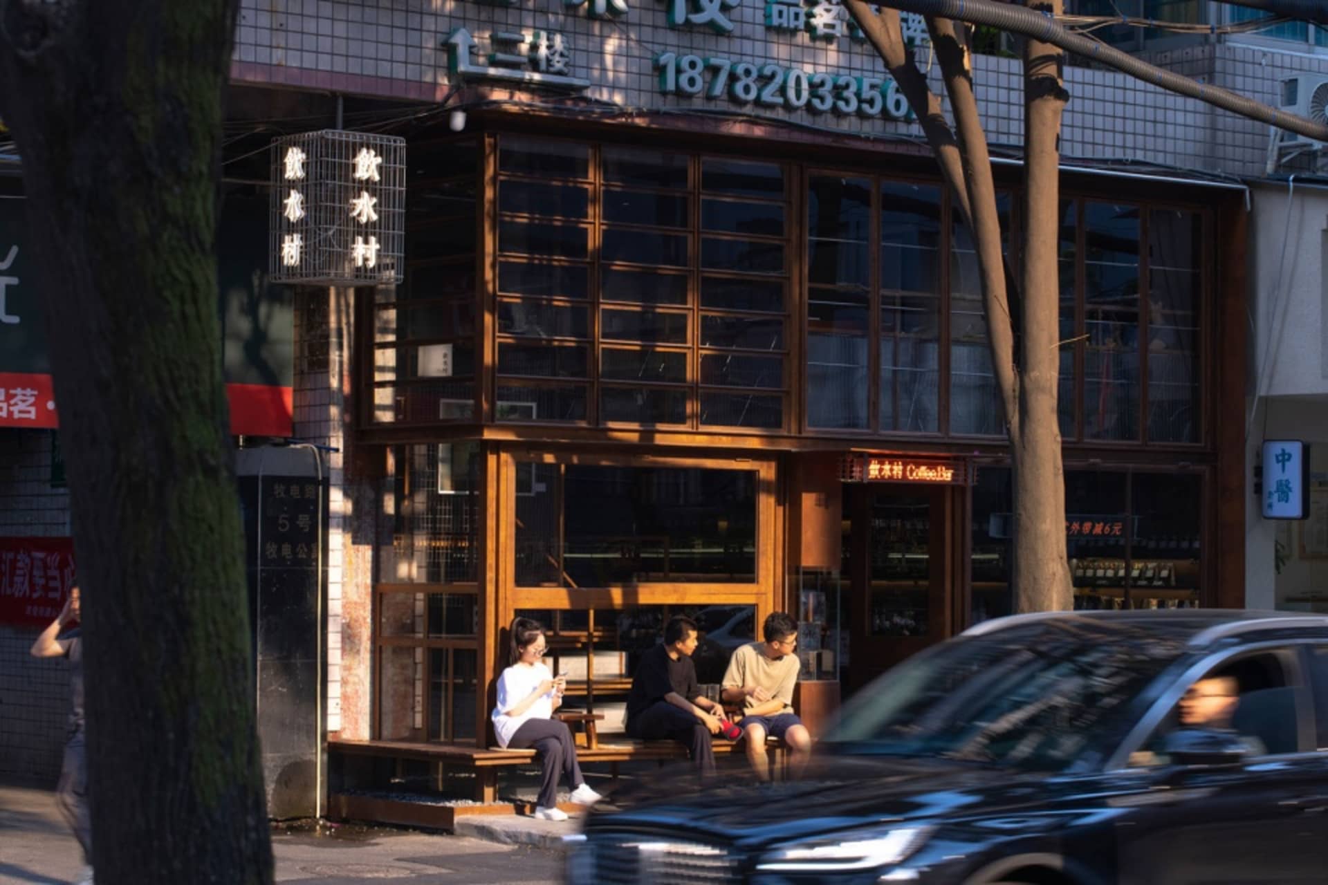 An Asian restaurant with people sitting outside on wooden benches in front of the establishment, surrounded by glass walls and natural light