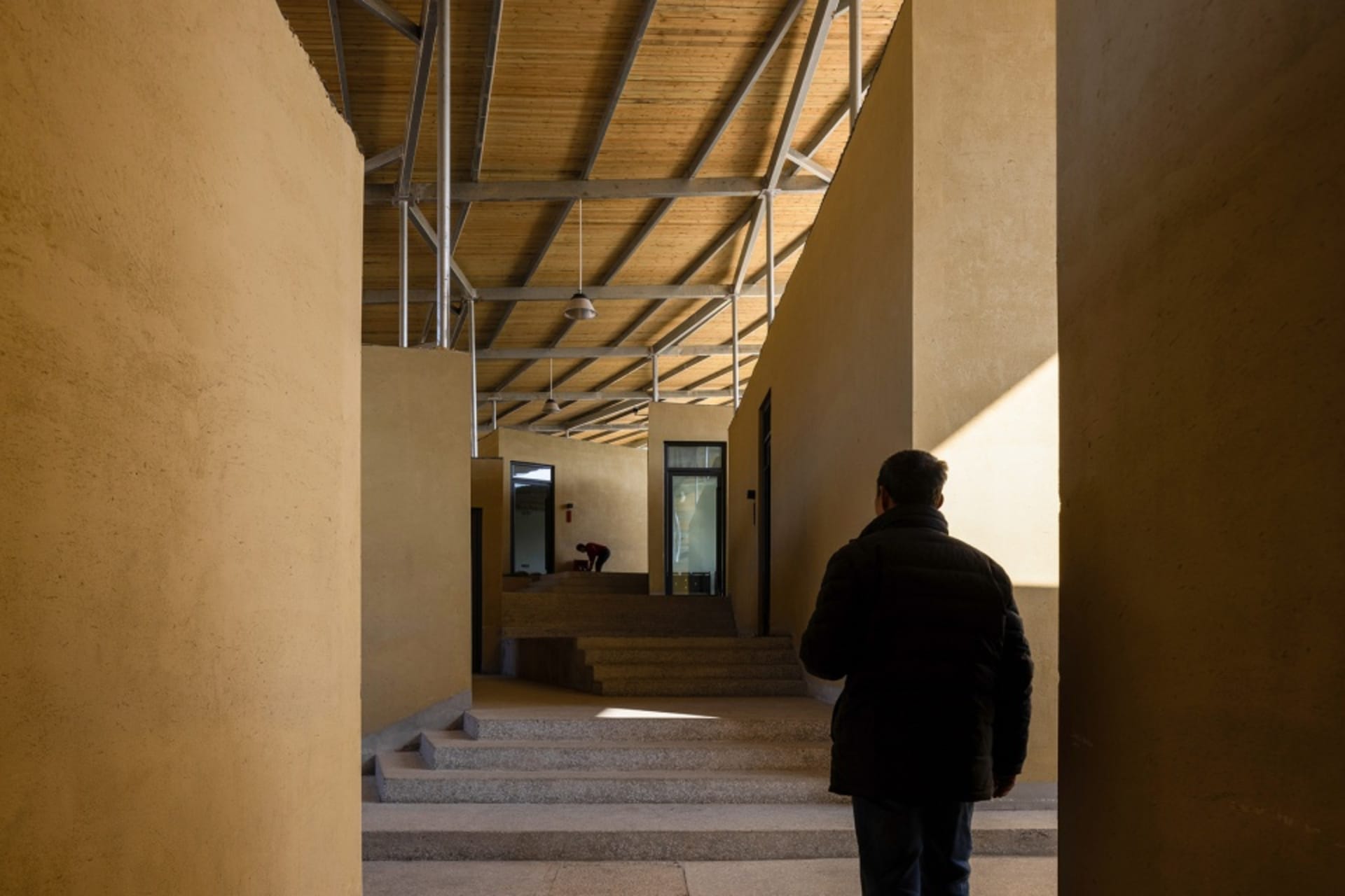 A hallway with concrete walls, featuring steps leading upwards and two people, one near the left side and one further back on the right side
