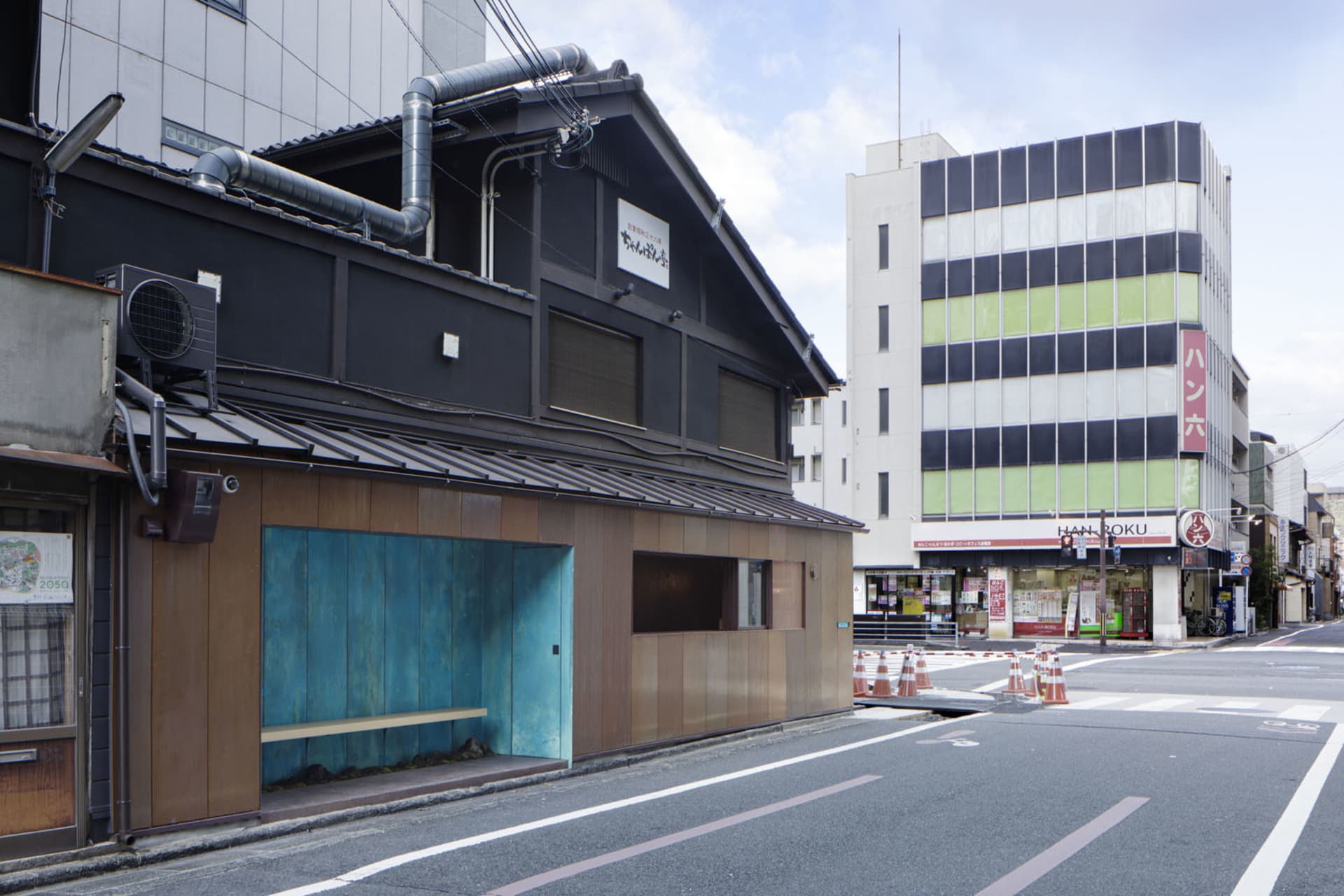 A modern building with a wooden facade and a blue roof, featuring a small shop or cafe in front, outdoor benches, and construction barriers