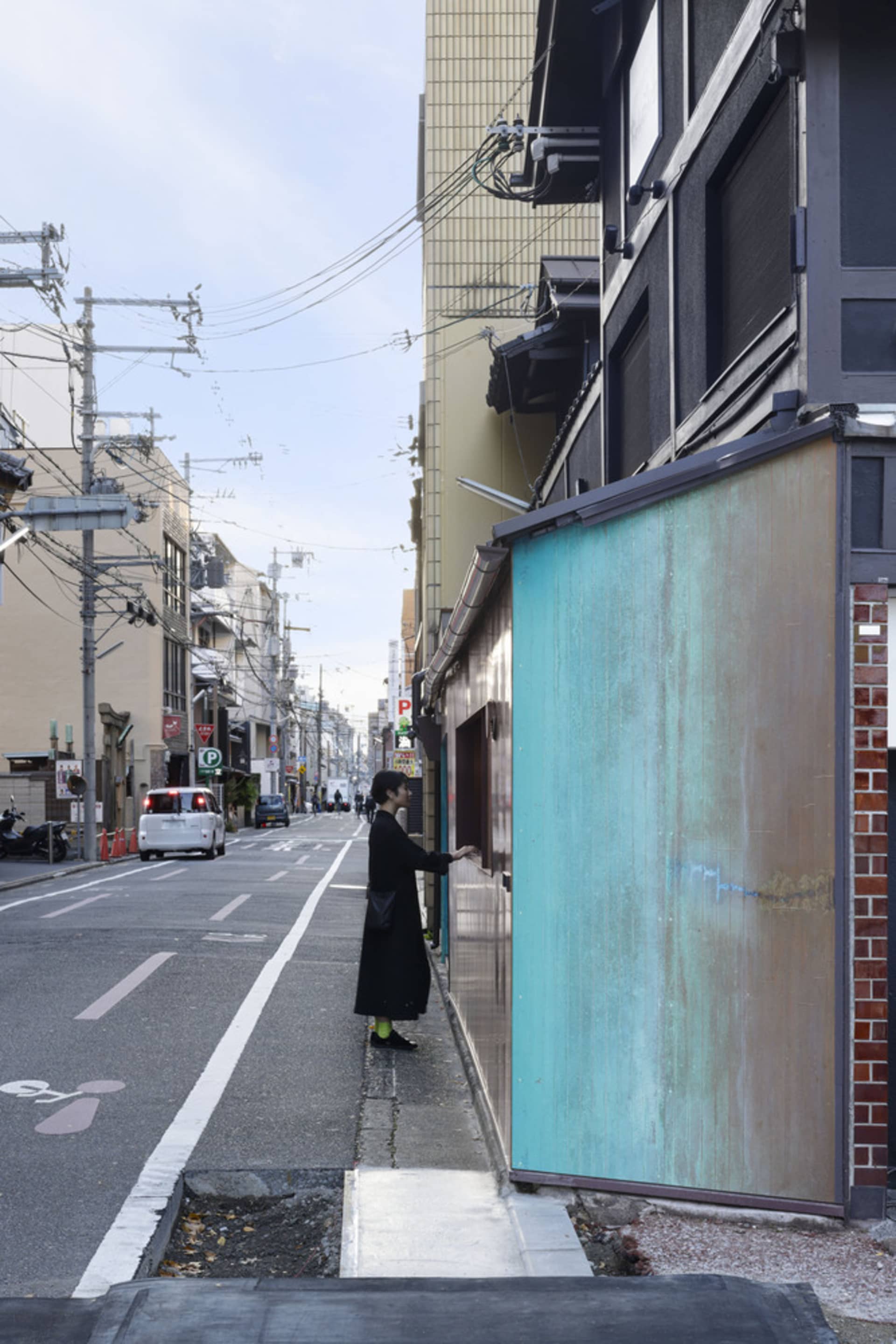 A woman stands on the sidewalk next to a building, looking at something in front of her, with several cars and a motorcycle parked nearby in a bustling city environment