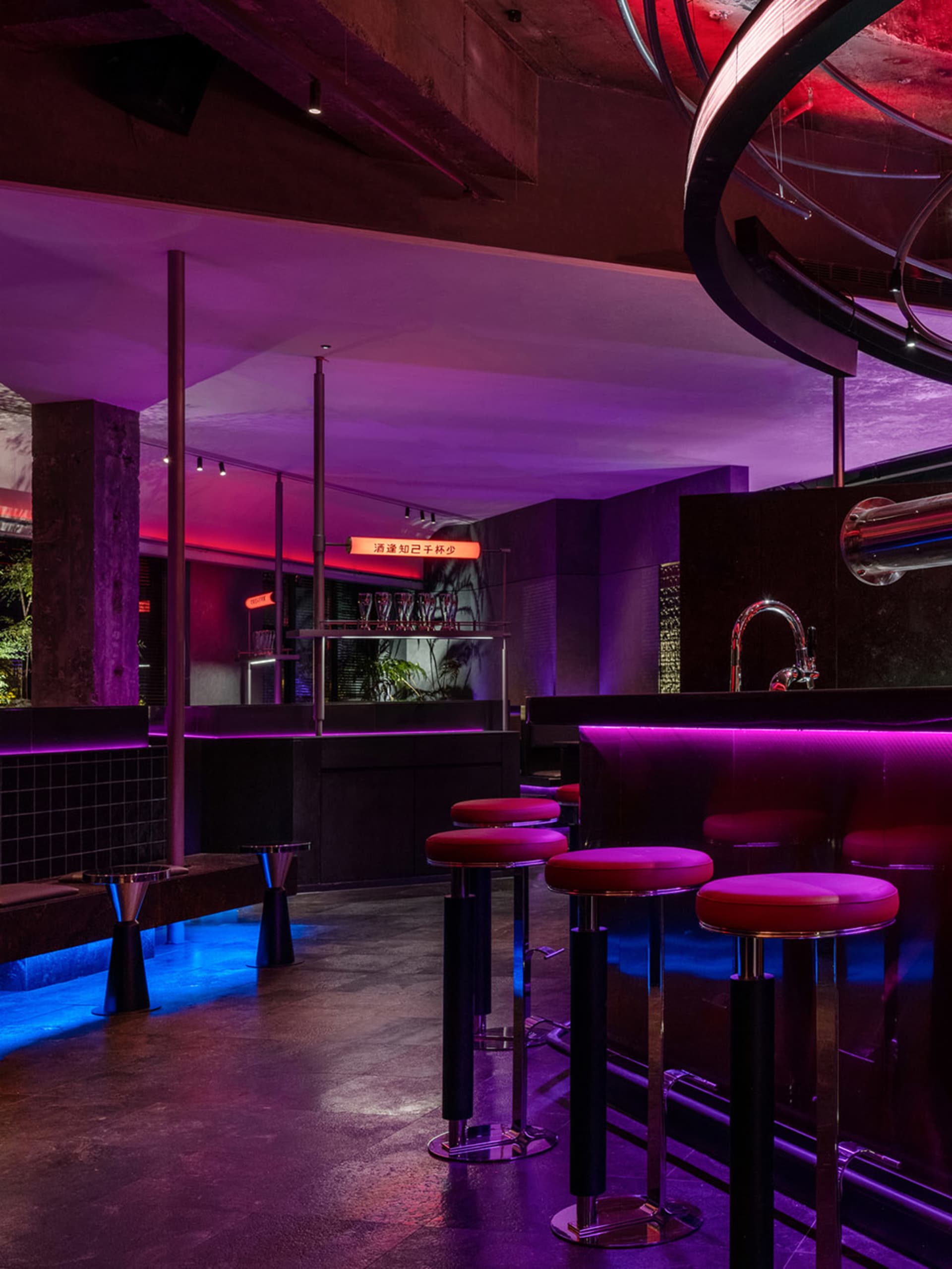 A modern bar area with purple neon lighting, chairs, and potted plants, creating a stylish and inviting atmosphere