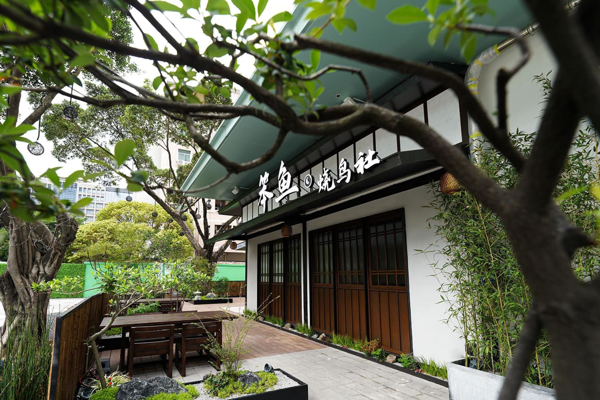An Asian restaurant with a wooden exterior and green roof, featuring a patio area with chairs and dining tables, surrounded by potted plants