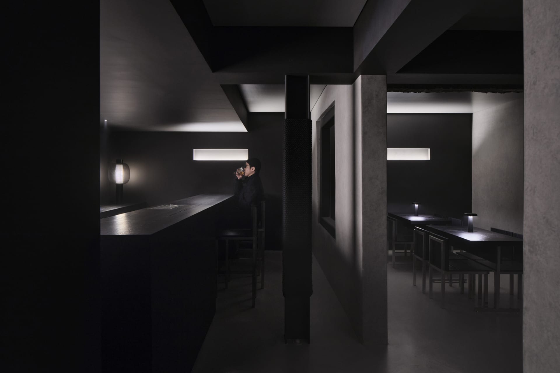 A dimly lit bar area with dark wood accents, multiple dining tables, and a bar counter with chairs, set in a restaurant
