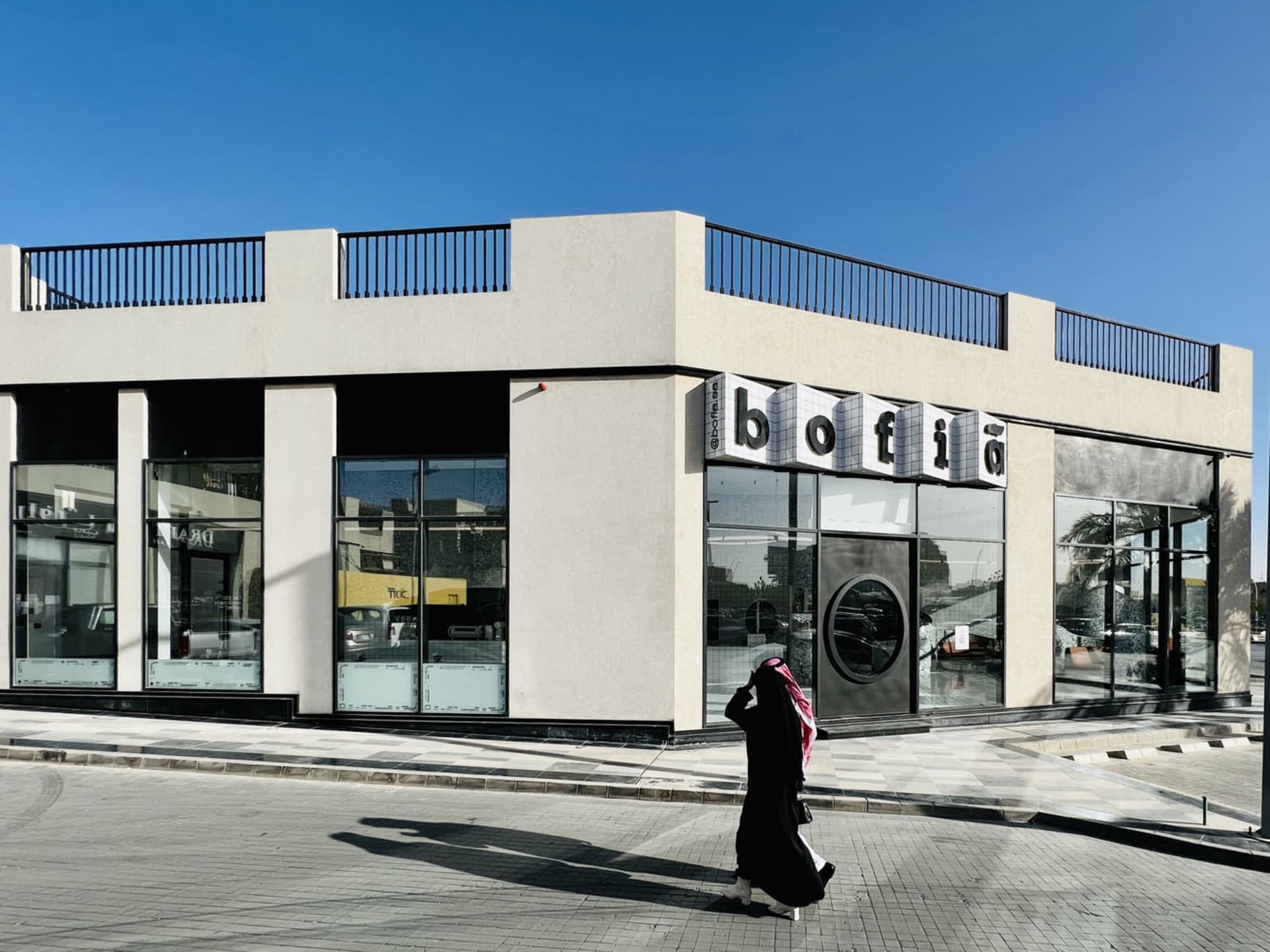 A casual street scene with a woman walking in front of a building, surrounded by parked cars, outdoor furniture, and a dining table, set against a relaxed urban backdrop