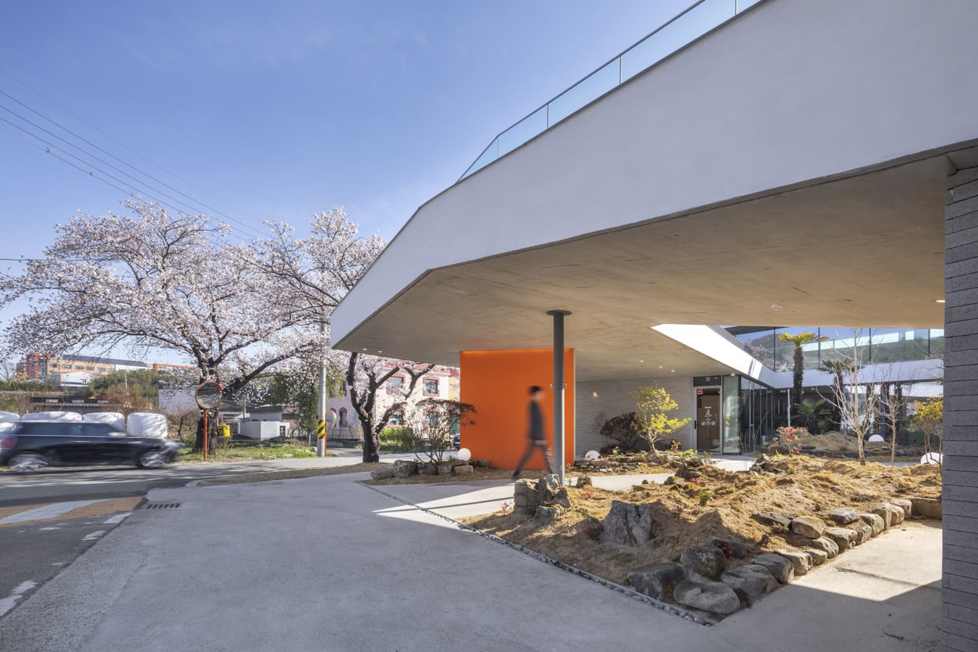 A modern building with a large glass wall extending over a walkway, surrounded by trees and rocks, with people walking around outside