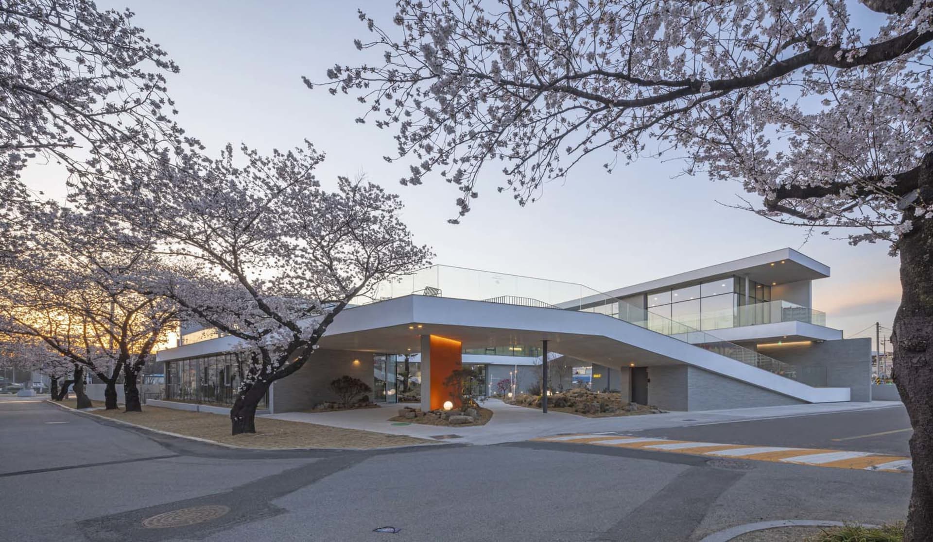 Modern building with a large glass wall, surrounded by trees and flowers, featuring a curved roof and a serene atmosphere