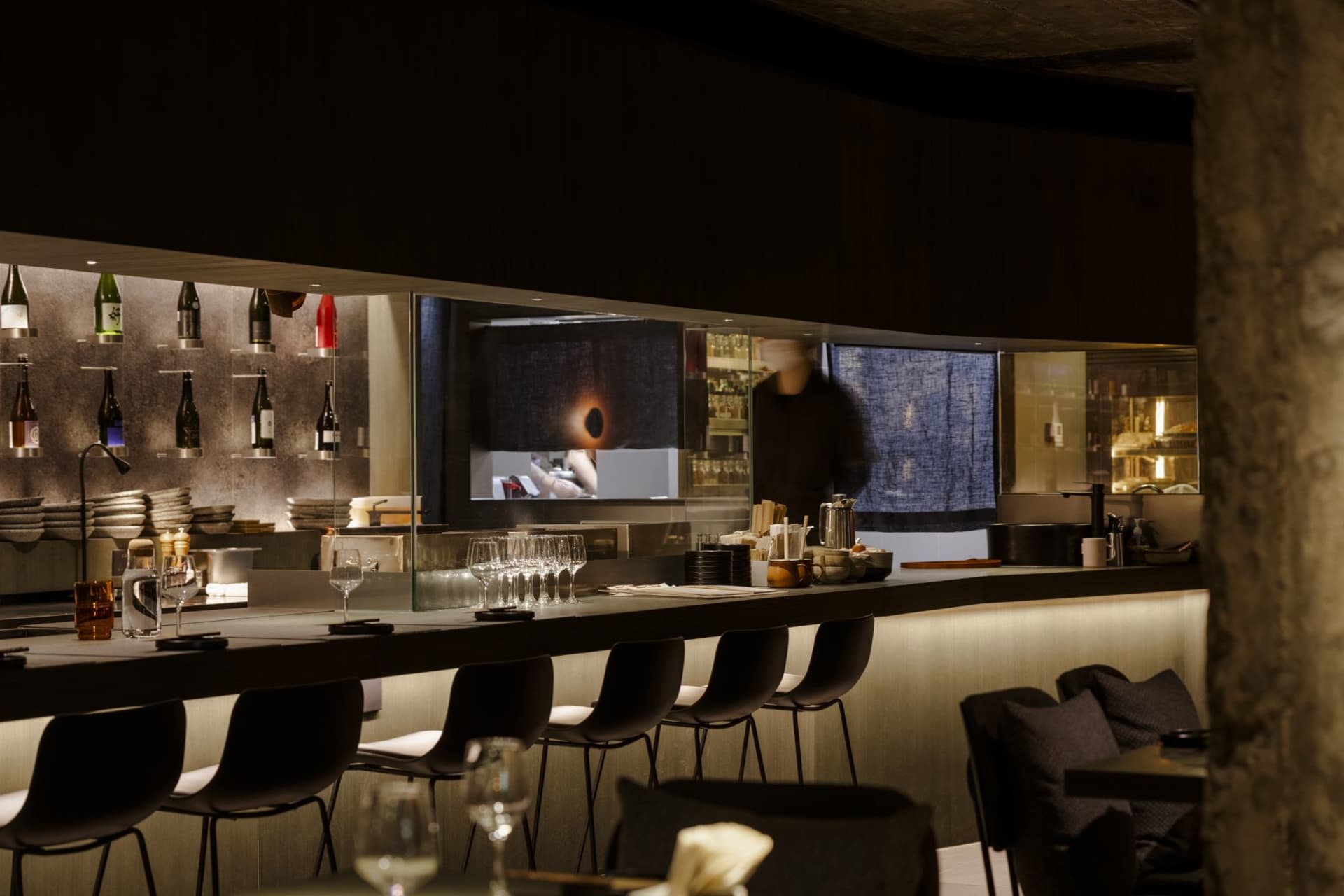 A restaurant bar scene with a counter, wine glasses, and chairs around a dining table, with scattered bottles and an inviting atmosphere