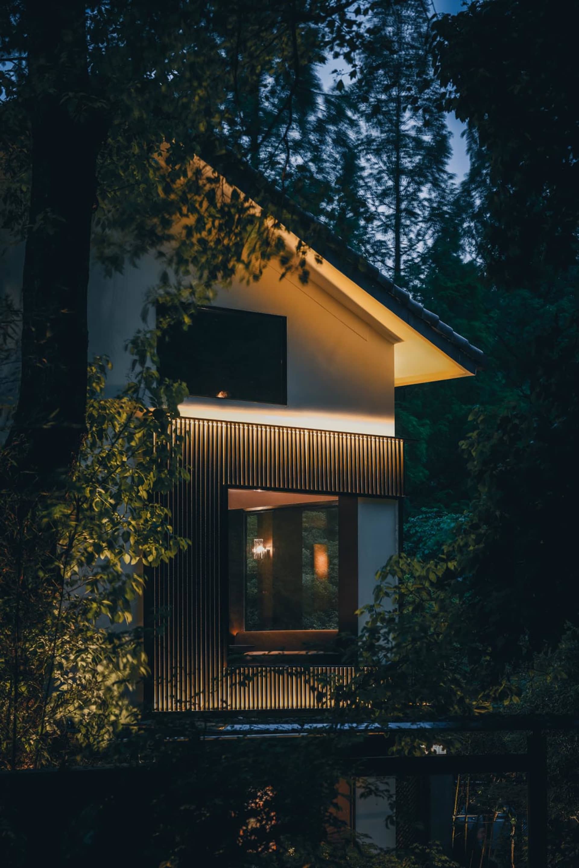 夜晚燈光明亮的現代住宅，附大型白色建築、木門與兩扇窗戶，室內可見椅子
