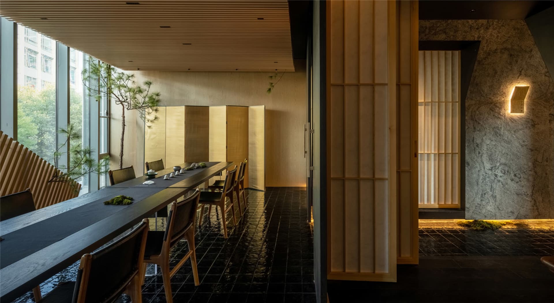 Cozy Asian restaurant dining area with a long wooden table and chairs, set with bowls, cups, and spoons, surrounded by modern wood elements and warm decor