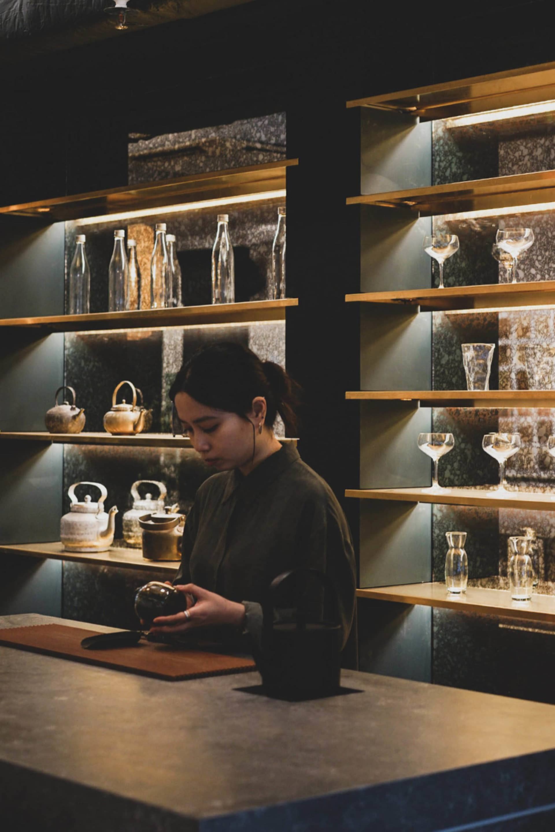一位女性站在擺滿酒杯、杯子與酒瓶的木質吧台前，周圍瀰漫著社交聚會的氛圍