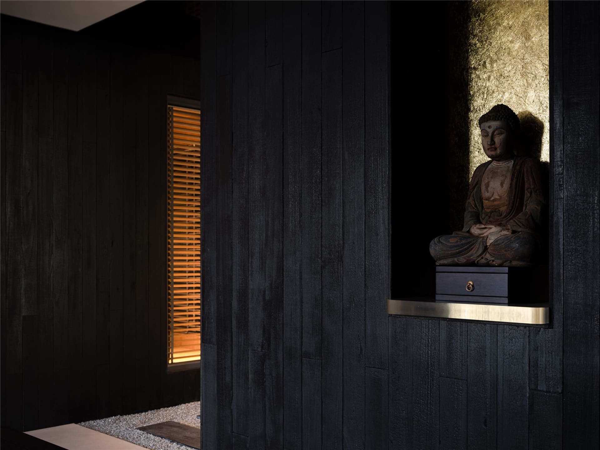 A dark room with a wooden Buddha statue on a pedestal, two windows, and a solemn atmosphere