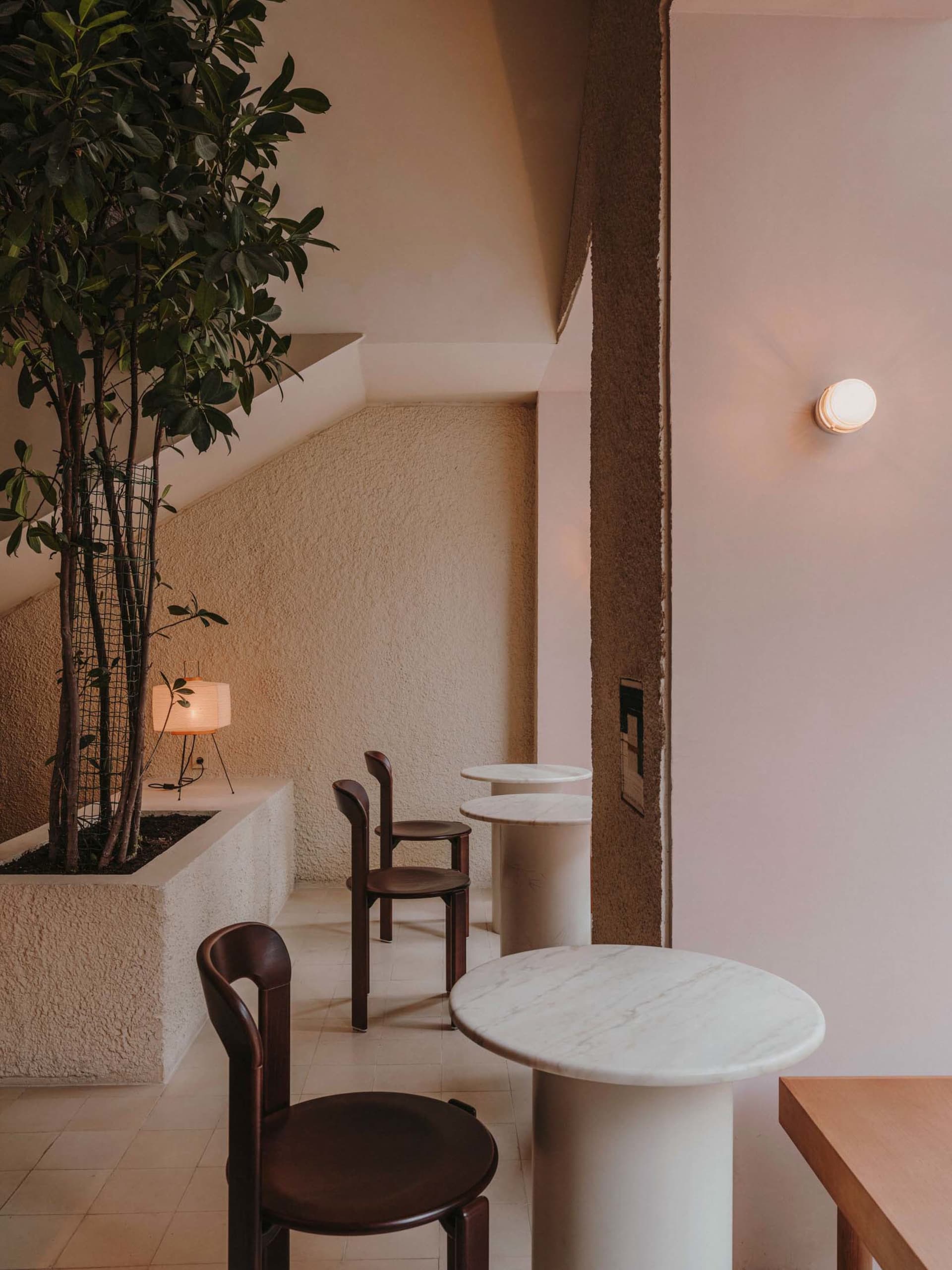 Cozy modern dining space with two white tables, three chairs, lamps, and a potted plant, set in a neutral-toned environment with clean lines