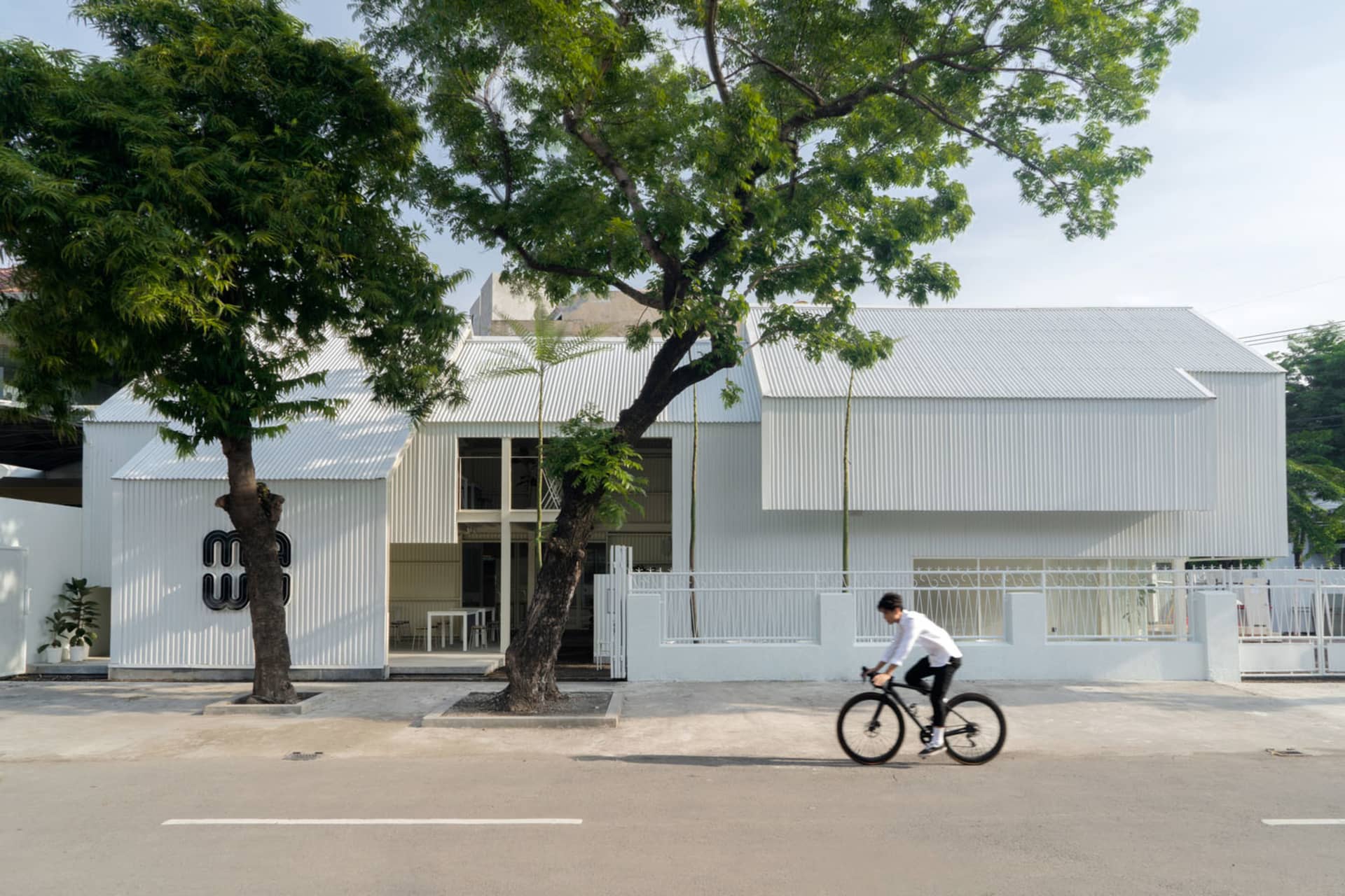 A modern building with a white concrete facade, featuring outdoor seating areas with two chairs and a bench, set in a calm and peaceful environment
