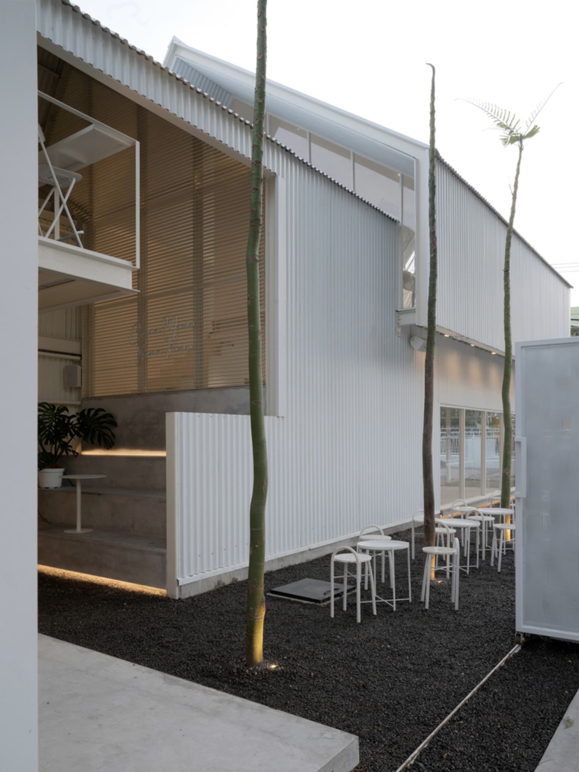 A modern building with a patio area featuring white chairs, dining tables, and potted plants, surrounded by concrete walls and natural light