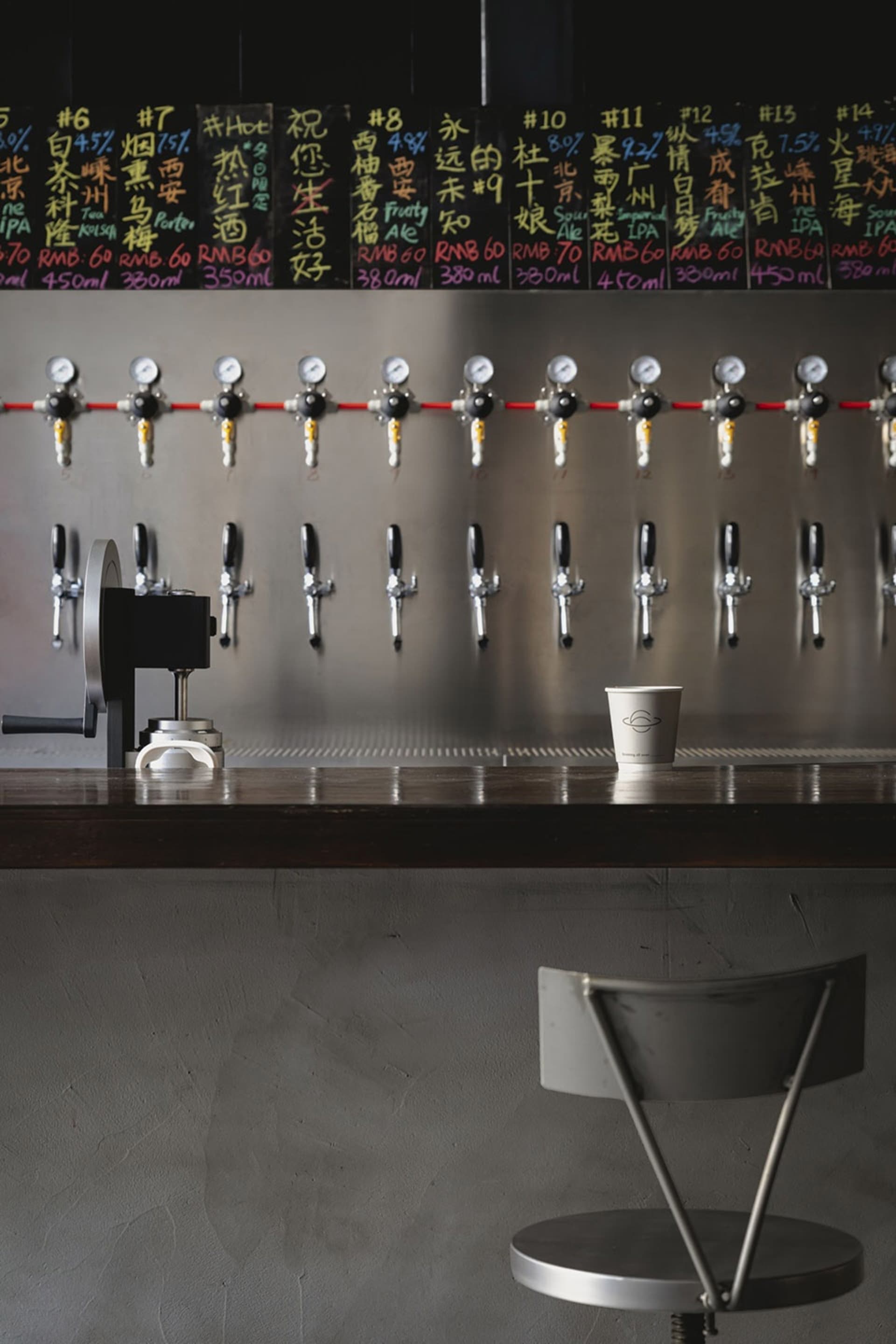Industrial-style bar area with a long counter featuring 14 beer taps and two cups, set in a busy restaurant or bar atmosphere