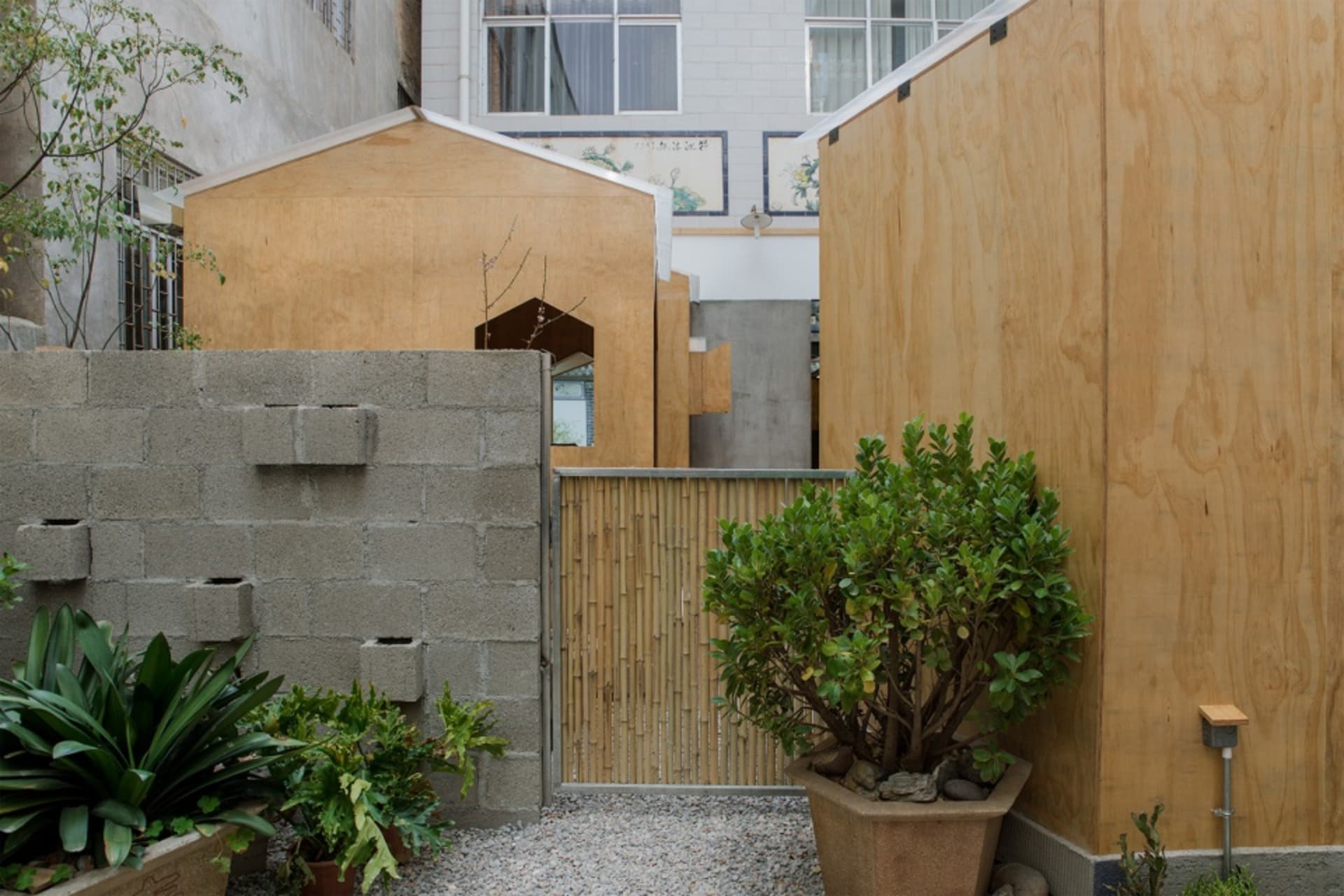 Two houses connected by a bridge, one with a wooden exterior and the other with a stone exterior, surrounded by potted plants and vases, with large windows allowing natural light in
