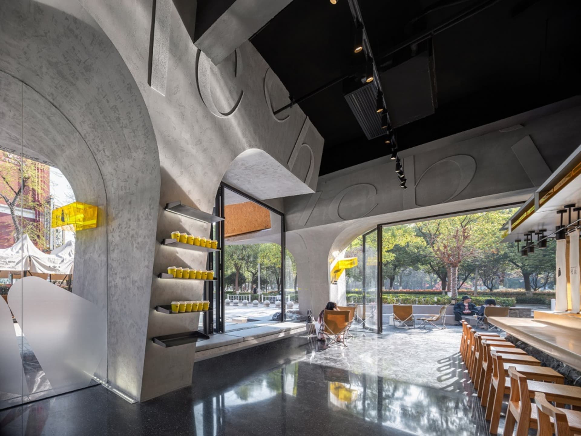 A modern restaurant interior with concrete walls, wooden elements, and large windows allowing natural light to enter, featuring dining tables and chairs