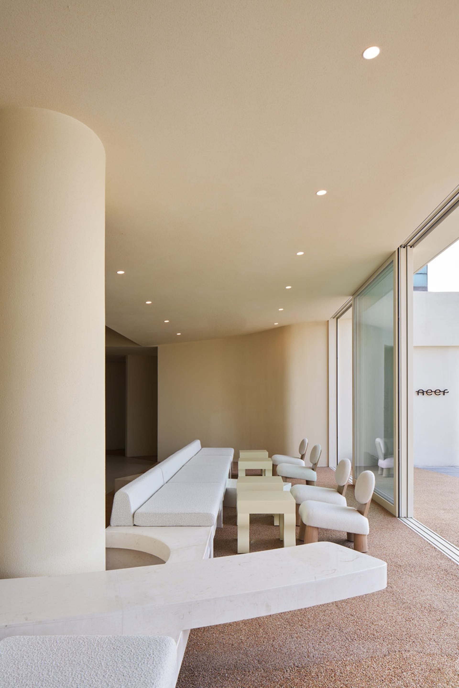 Modern open space with a glass wall, a long bench, and multiple chairs, surrounded by white walls and furniture, with natural light filling the room