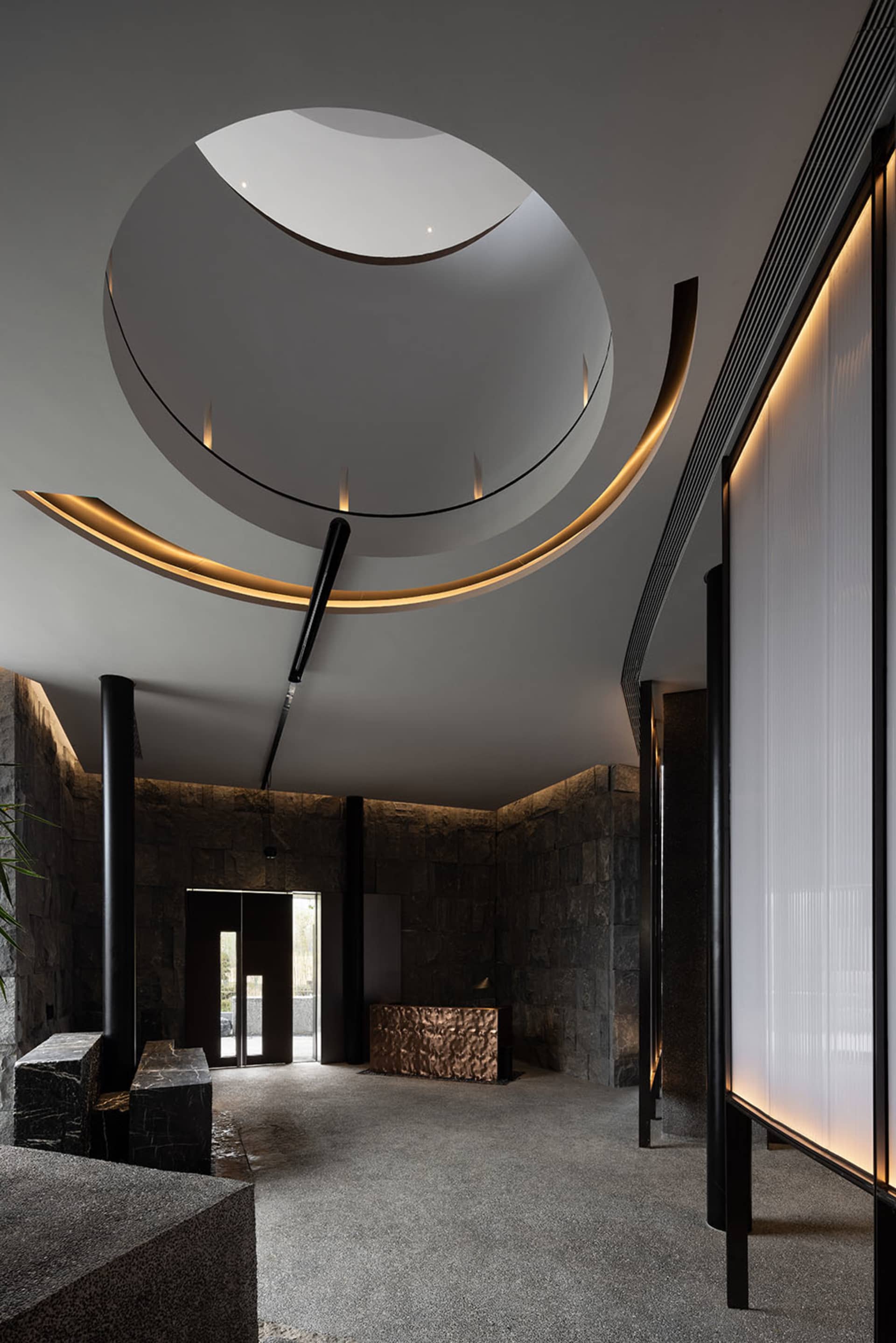 Well-designed hotel lobby with a stone wall, black doorway, and natural light from a circular ceiling window, featuring a bench and potted plants