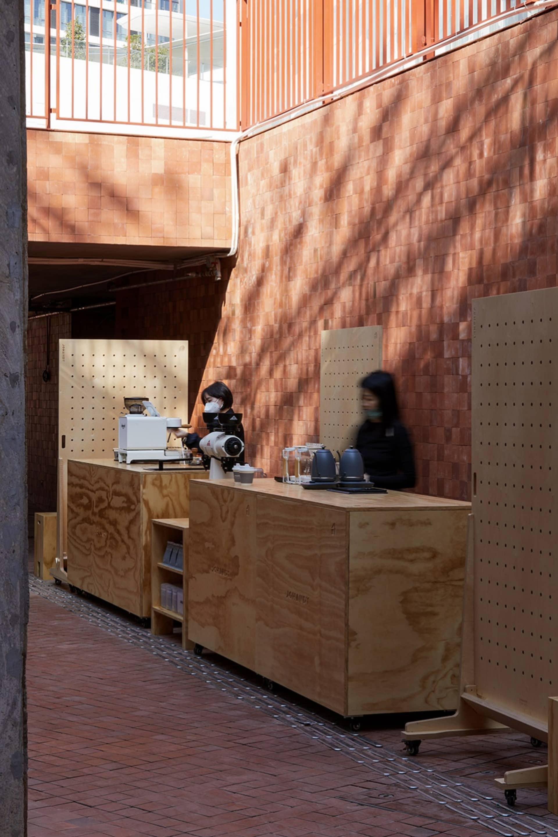 A casual cafe interior with a counter and seating area, featuring a woman wearing a mask and serving drinks and food