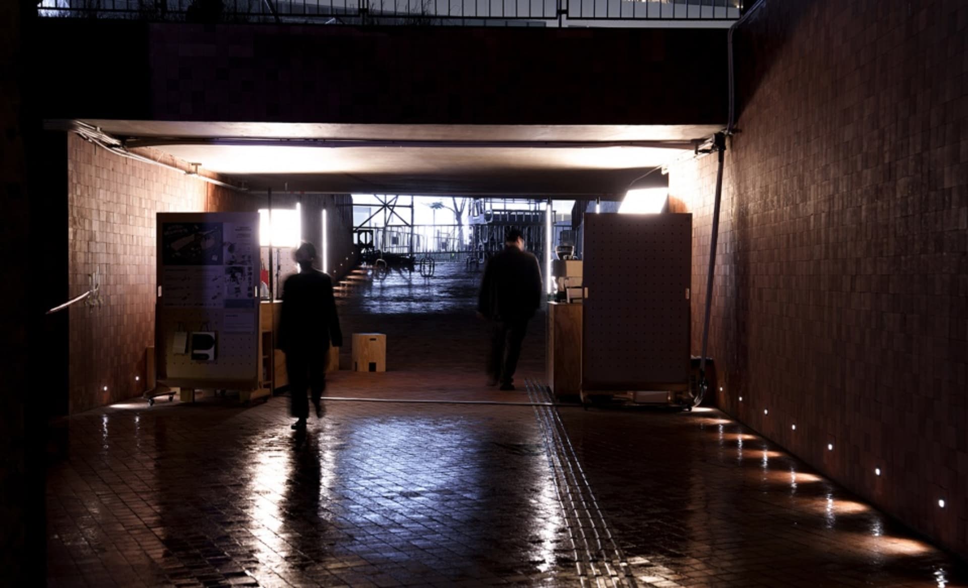 A brick-lined hallway with chairs and two people walking down the corridor, carrying boxes