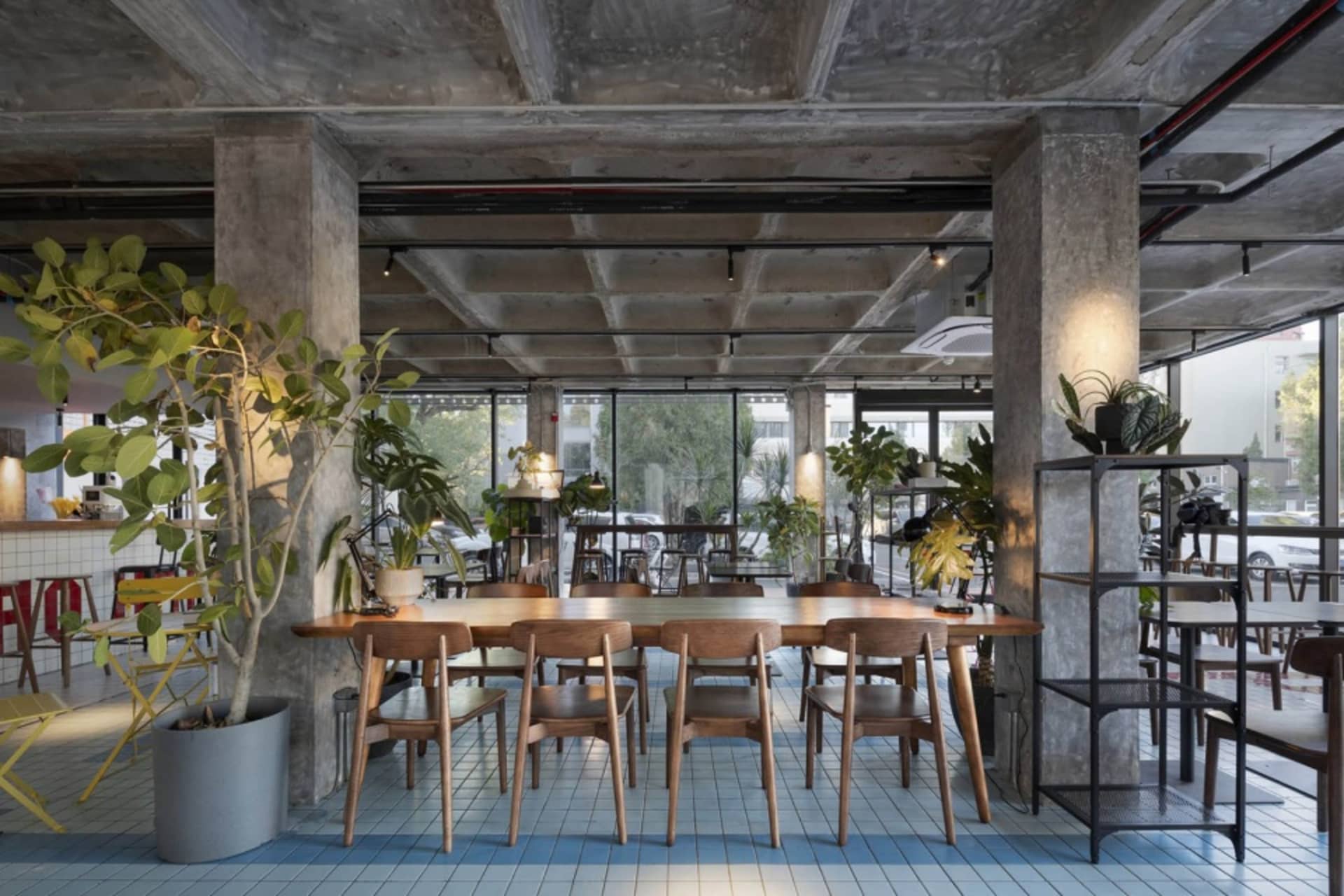 Modern restaurant interior with a large wooden dining table surrounded by metal chairs, potted plants, and vases, under natural light