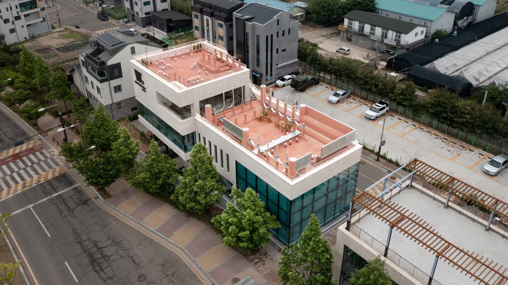 Modern building with a red roof, featuring a large side terrace, multiple levels, and a rooftop view of the city, with cars parked in front