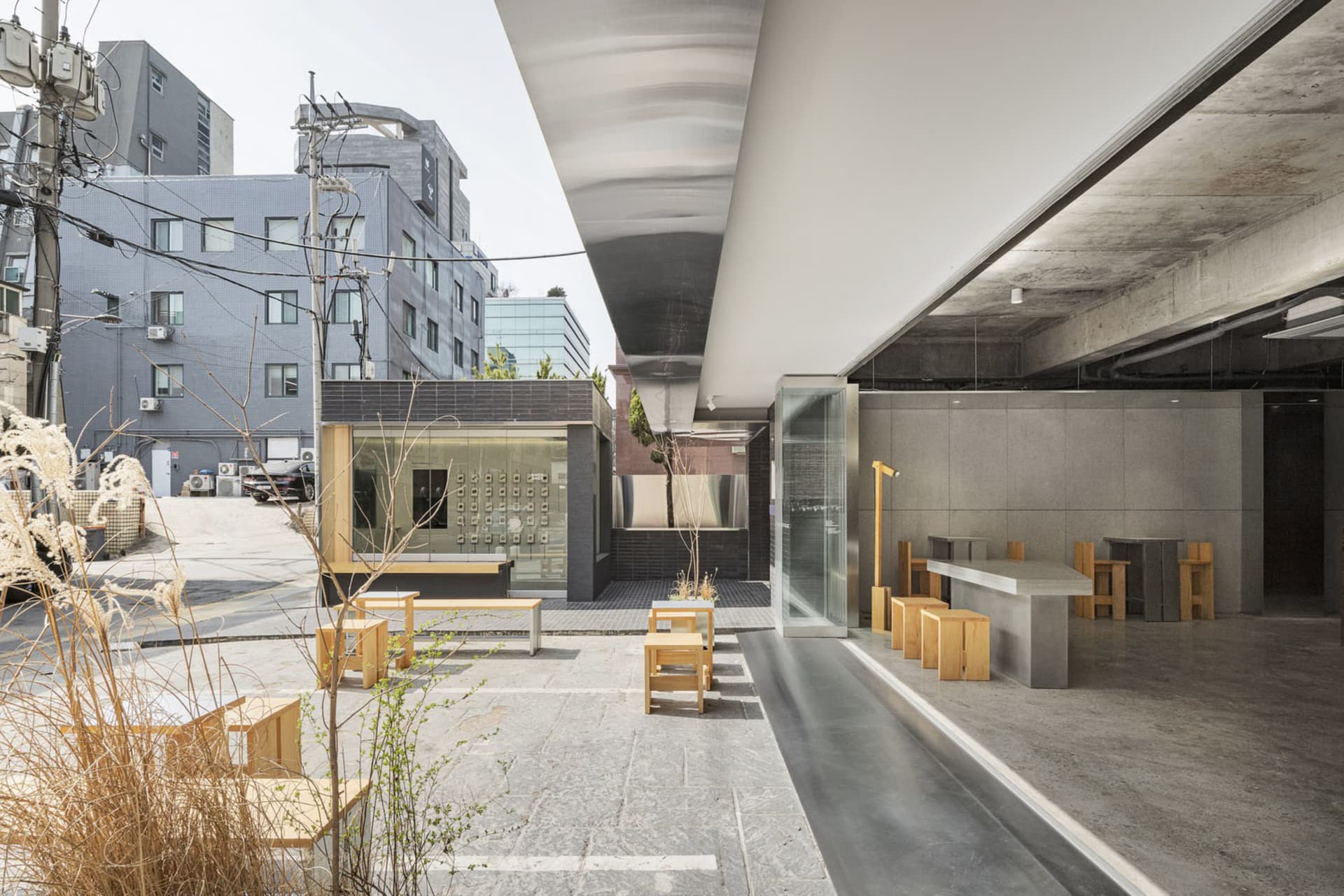 A modern building with a glass wall facade, wood and concrete elements, outdoor seating, and potted plants