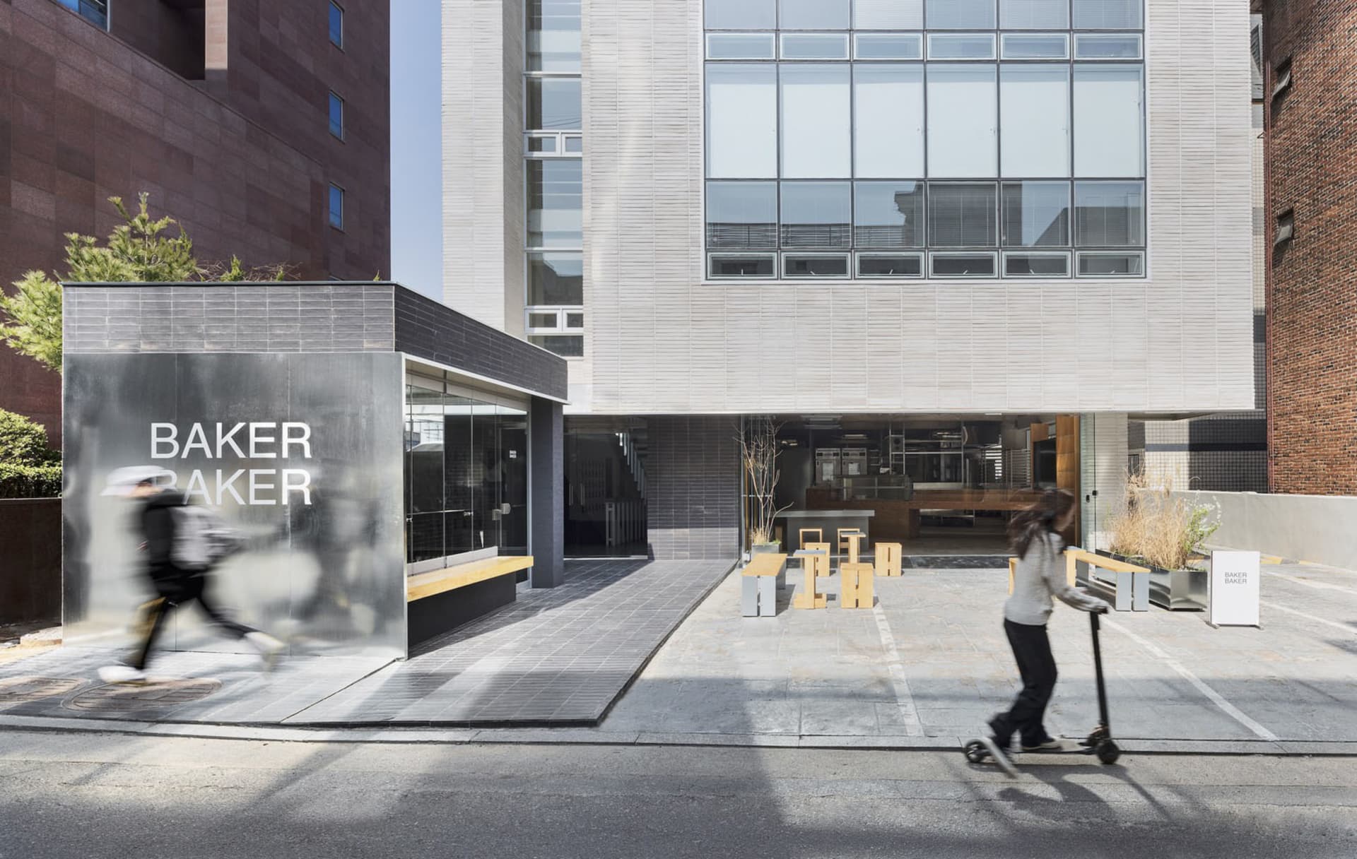 A modern building with a large window on its side, surrounded by benches and pedestrians, with one person walking and another on a skateboard