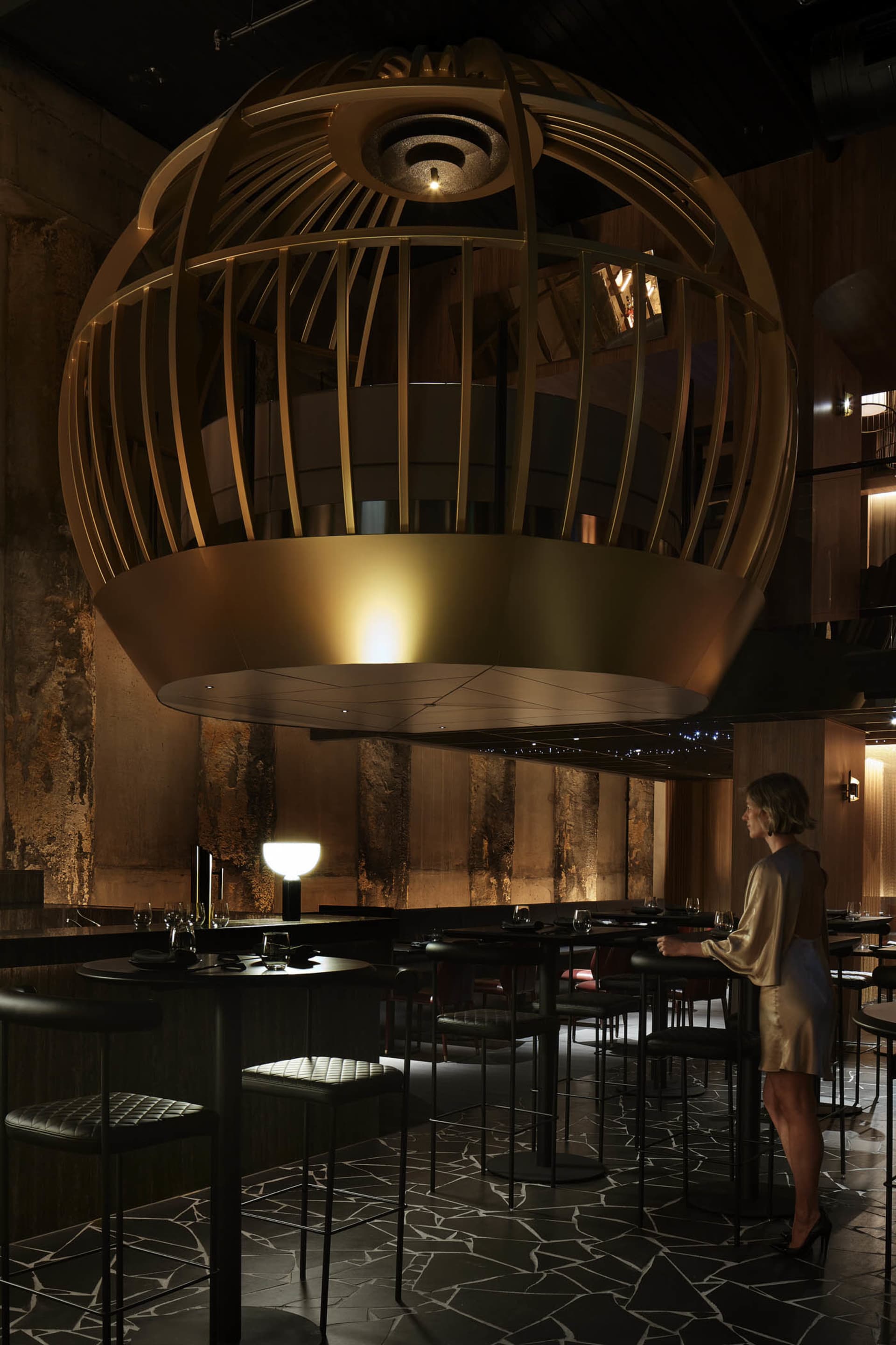 Dimly lit modern bar area with a dining table, counter, stools, and a golden sphere hanging from the ceiling