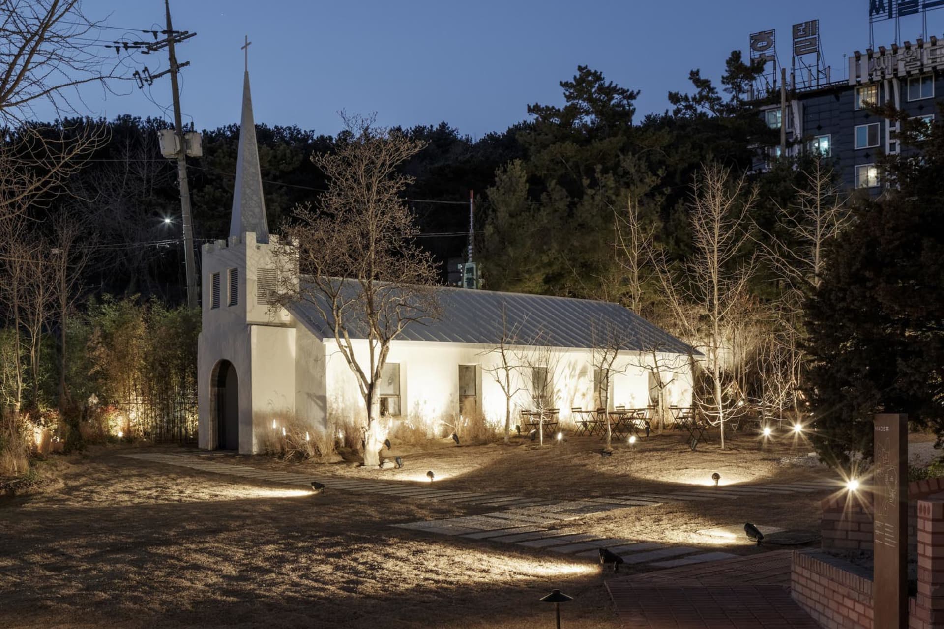 A serene old church at night surrounded by trees, with a white exterior and a cross on its roof, featuring benches for seating