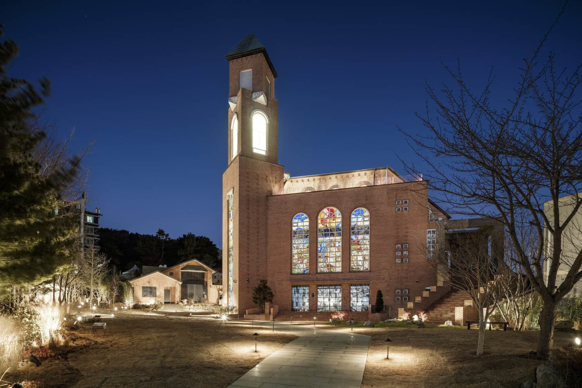 A large brick church at night with a clock tower, illuminated windows, and benches in the foreground
