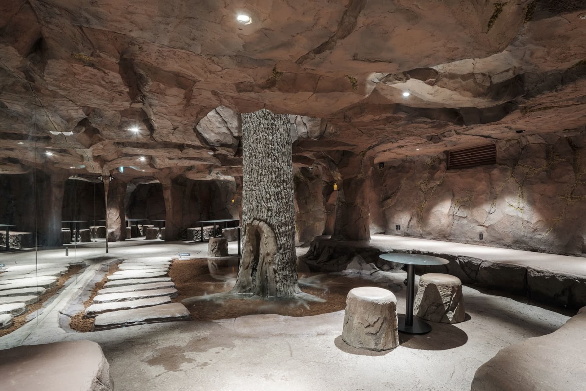 Cave-like cafe with stone walls, a dirt floor, and a large tree in the center, surrounded by chairs and dining tables