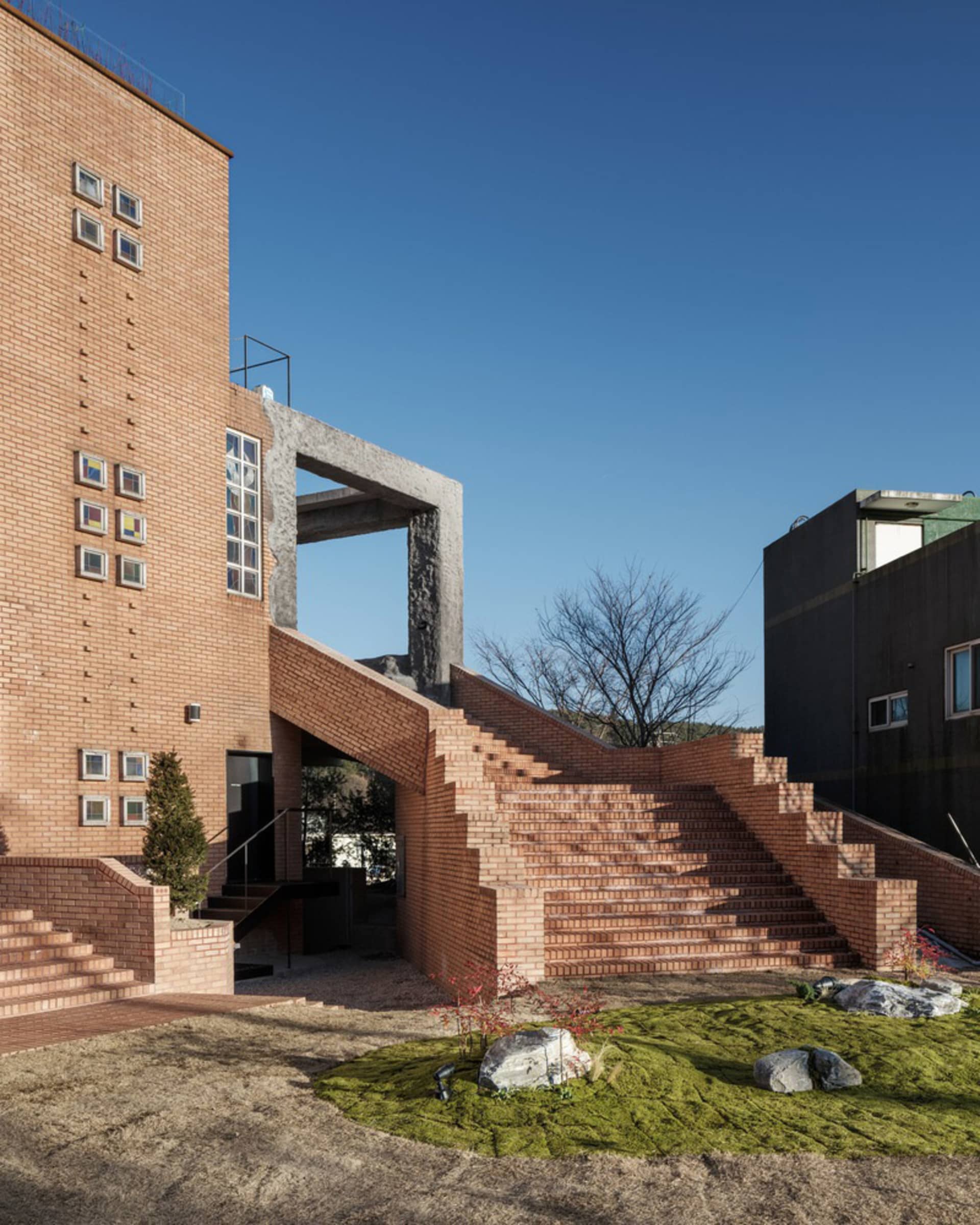 A brick building with multiple levels, featuring a mix of red bricks and concrete elements, with stone stairs and windows on various levels allowing for natural light